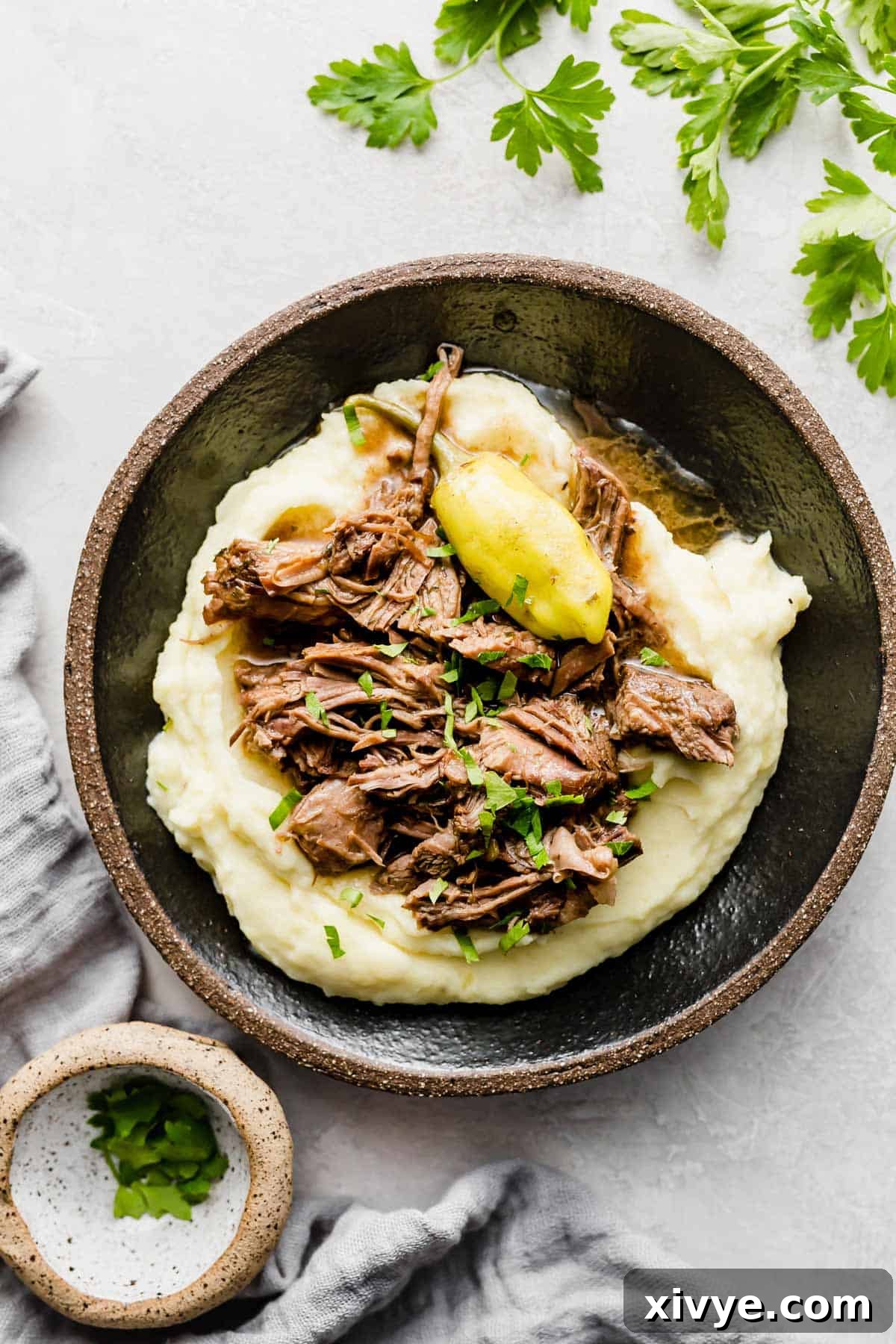 A black plate showcasing shredded Mississippi Pot Roast served over a bed of fluffy white mashed potatoes, garnished with green pepperoncini.