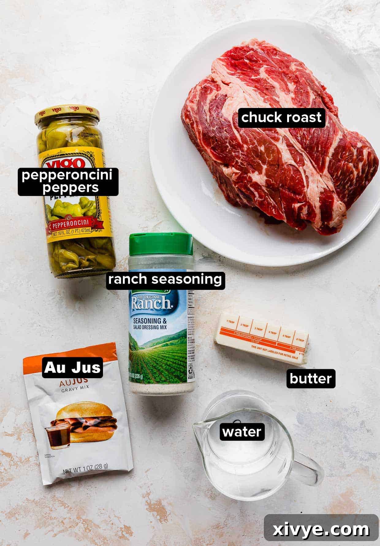 All ingredients for Mississippi Pot Roast laid out on a white background: beef chuck roast, jarred pepperoncini peppers, a packet of dry ranch seasoning, butter, water, and a packet of au jus seasoning.