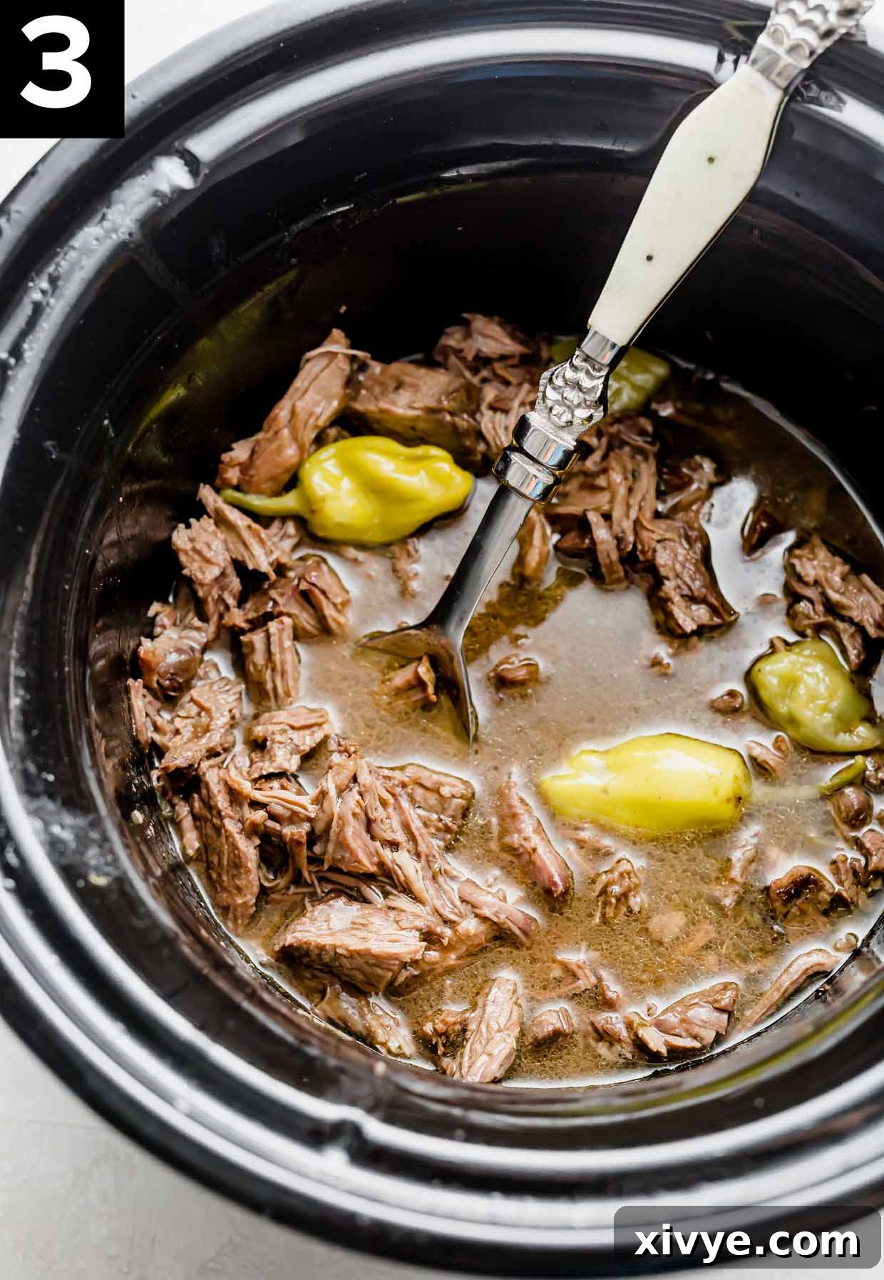 Close-up shot of shredded Crock-Pot Mississippi Pot Roast inside a black slow cooker, with visible pepperoncini peppers and rich gravy.