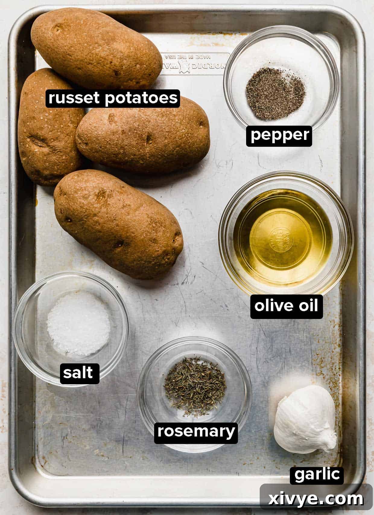 A baking sheet with salt, russet potatoes, garlic, olive oil, garlic, and rosemary on it.