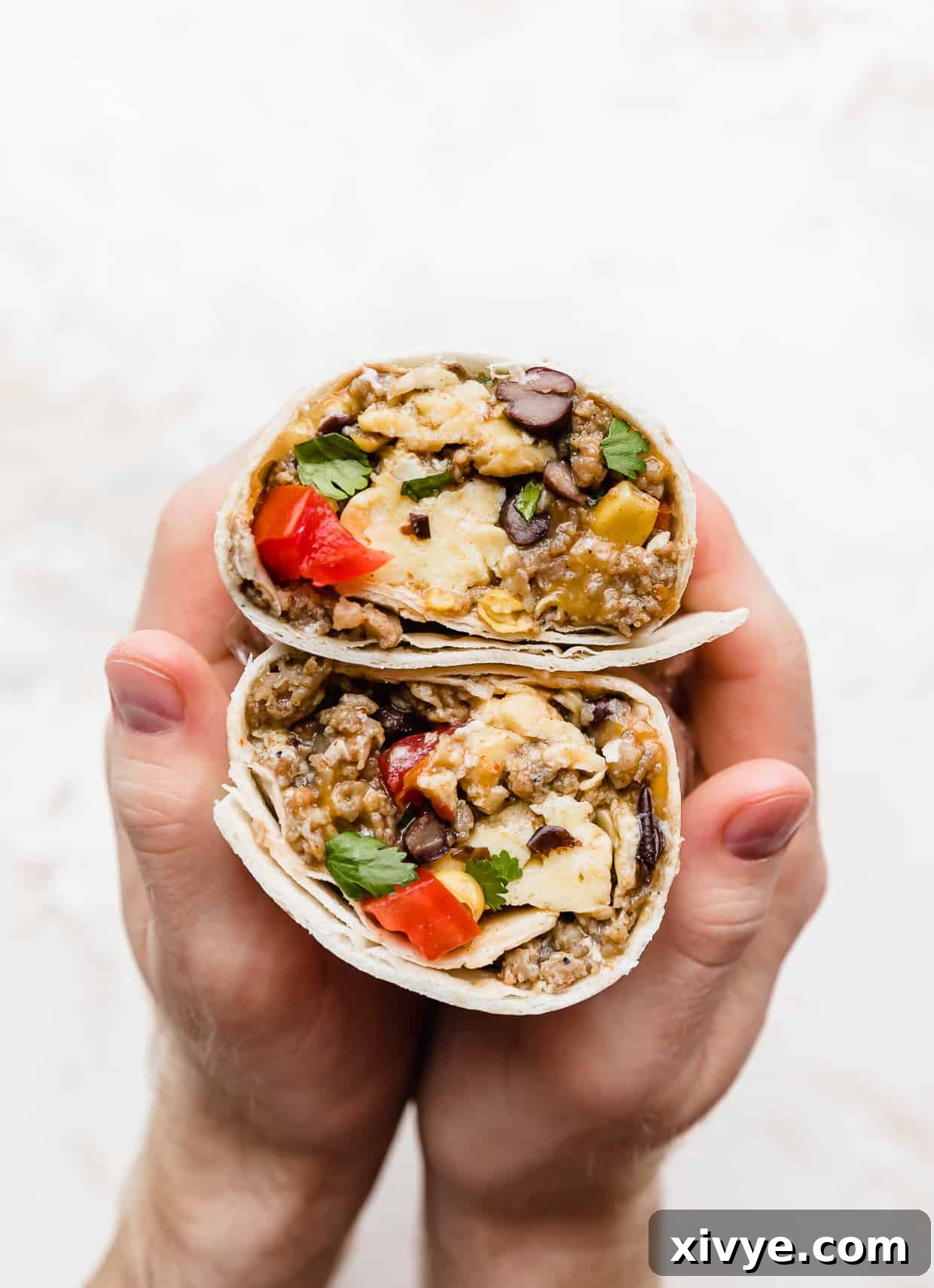 A set of hands holding a Breakfast Burrito with Black Beans and tomatoes that has been cut in half, to show the inside.