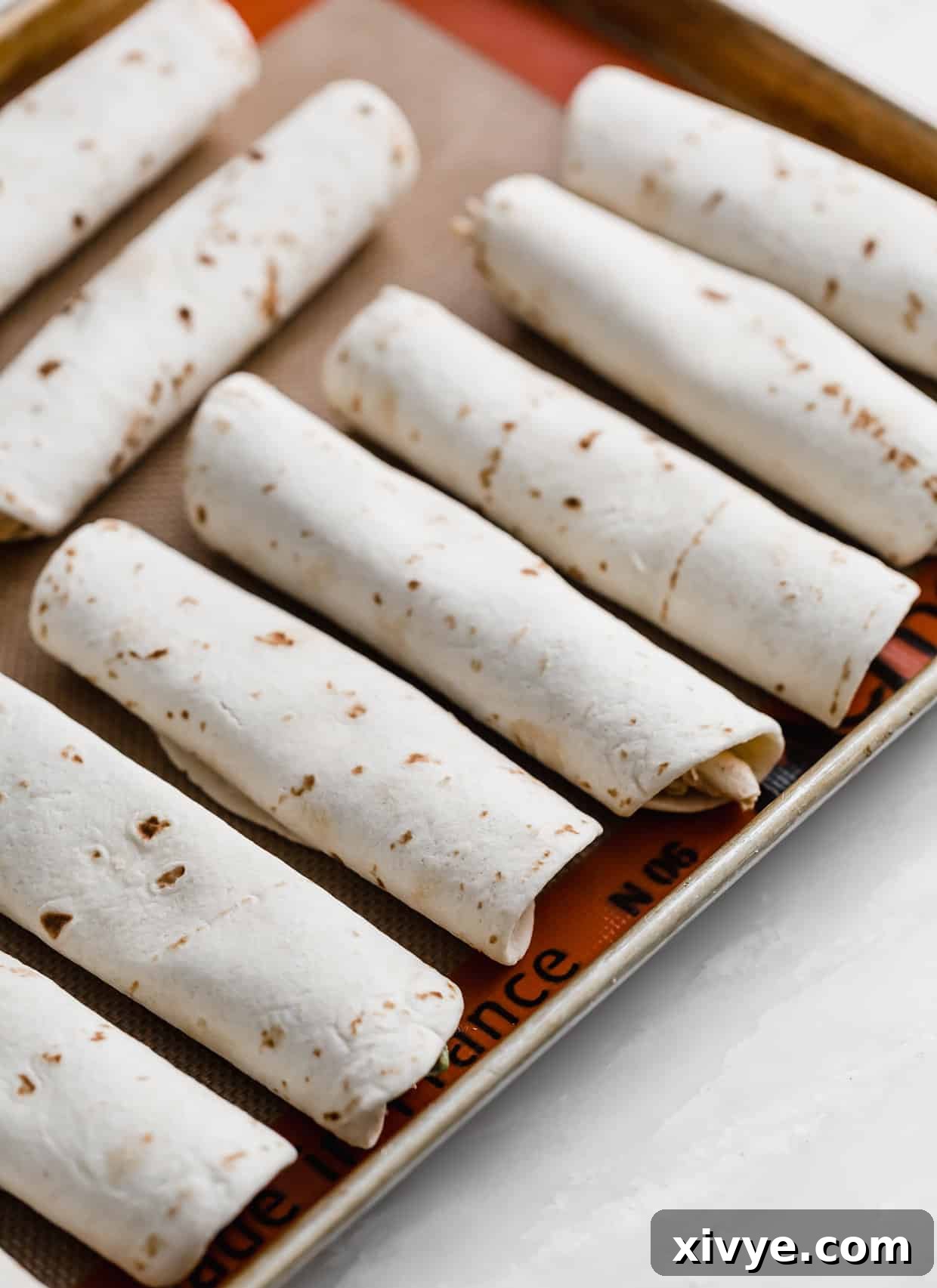 Cream cheese and chicken filled flour tortillas lined up on a baking sheet, ready for the oven.