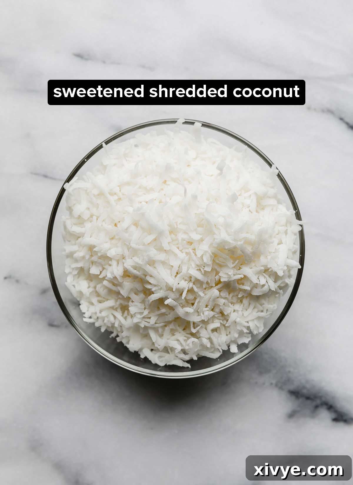 Sweetened shredded coconut in a glass bowl on a white marble background, ready for toasting.