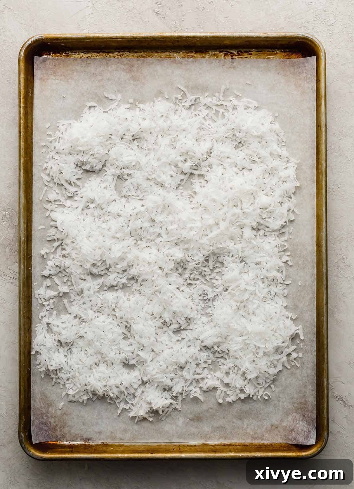 White sweetened shredded coconut spread evenly on a parchment-lined baking sheet, ready for the oven.
