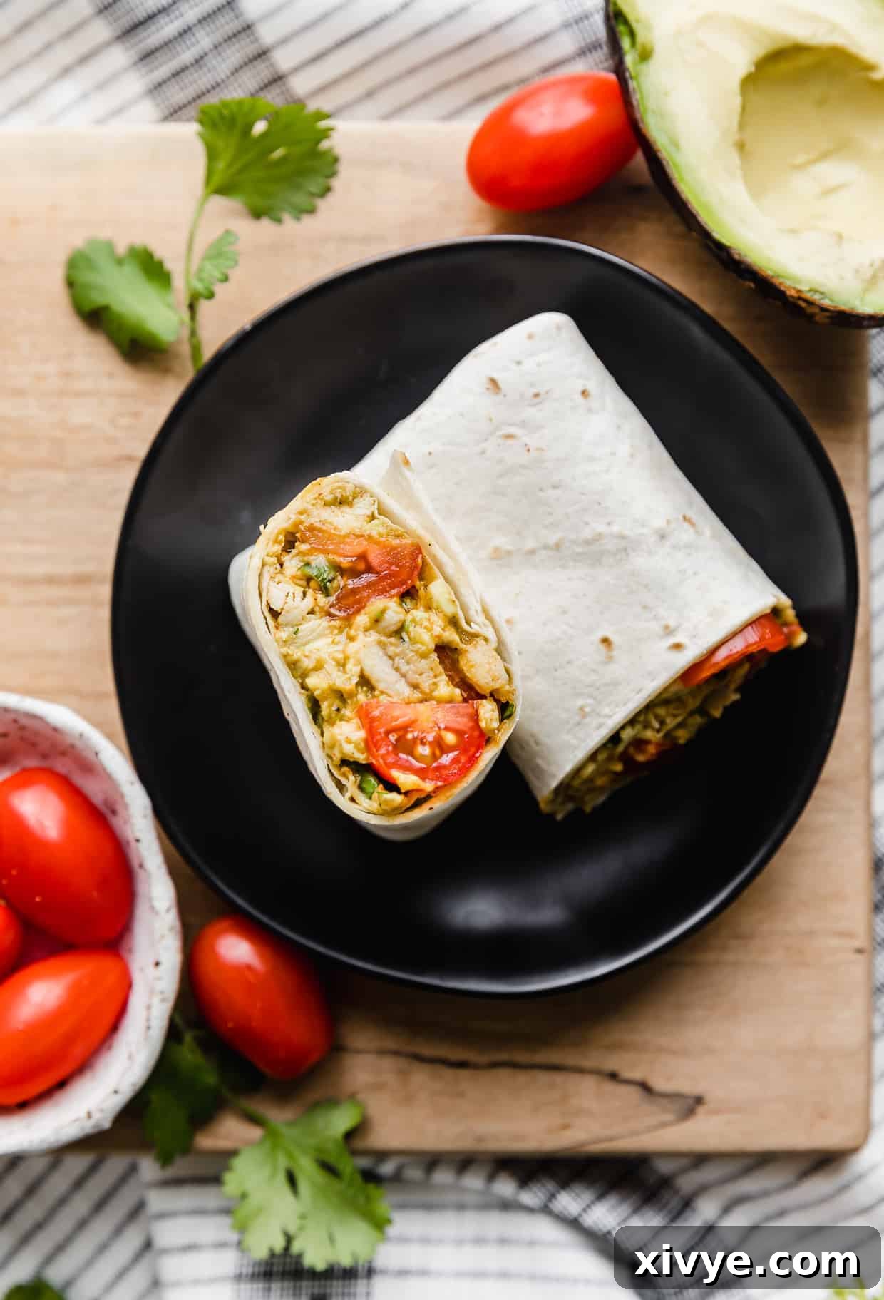 Creamy Avocado Chicken Wraps 3 A single avocado chicken wrap on a black plate, surrounded by whole grape tomatoes and sliced avocado.