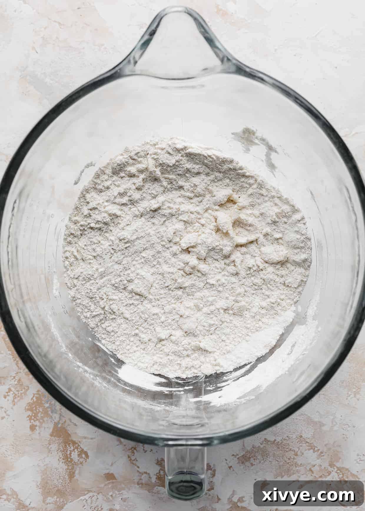 A clear glass bowl showcasing a finely sifted flour mixture, gently interspersed with small clumps of softened butter, prepped for mixing.