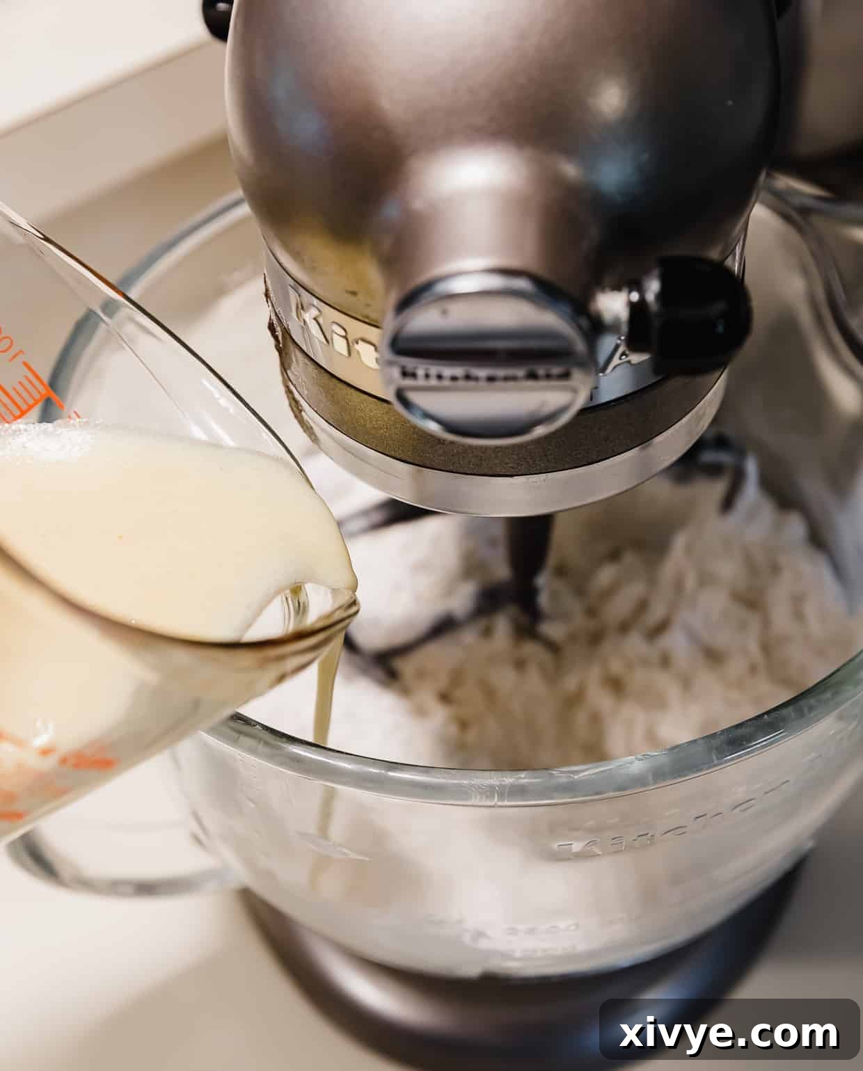A steady stream of liquid ingredients pouring into a stand mixer bowl containing the dry mixture, beginning the batter creation.