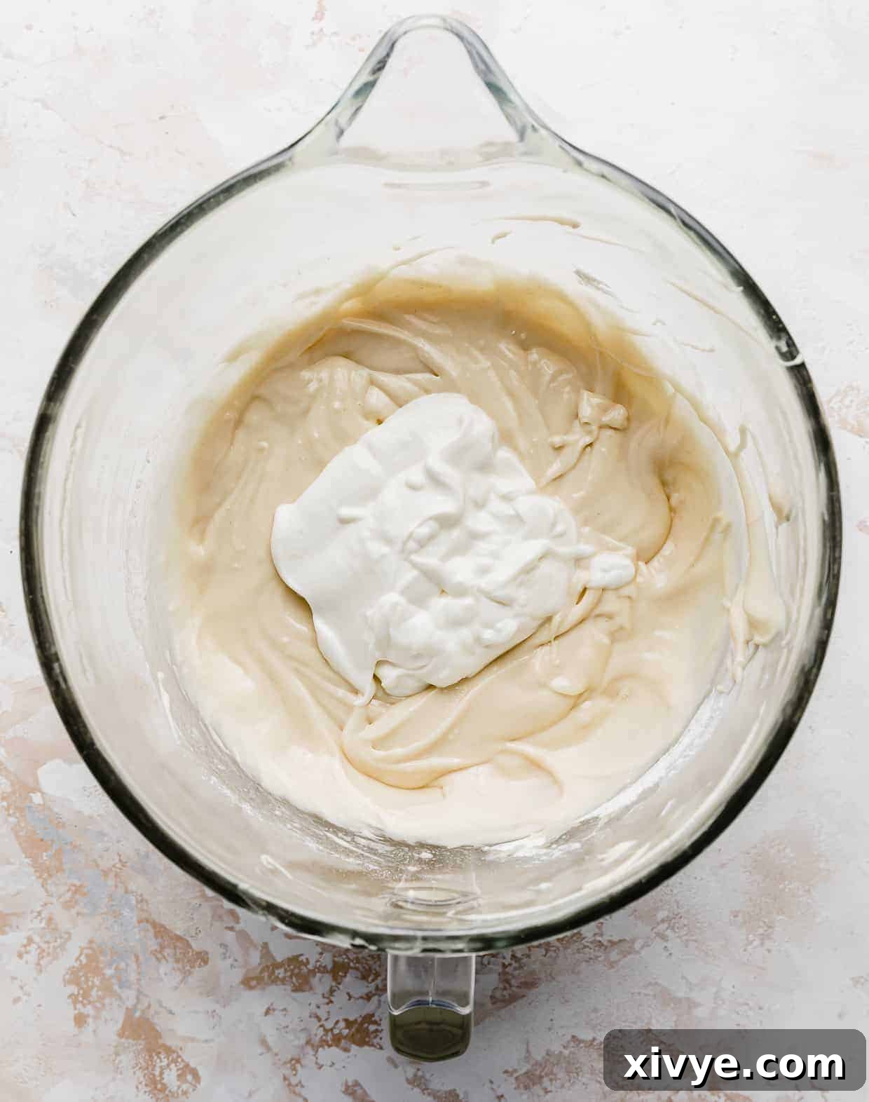 A dollop of rich sour cream positioned atop the smooth cake batter within a glass mixing bowl, awaiting incorporation.