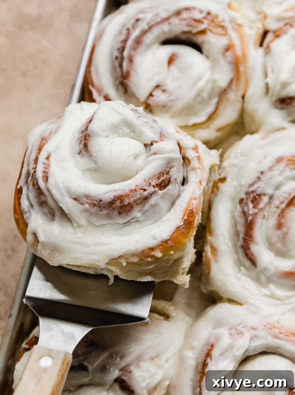 A large bakery-style cinnamon roll on a metal spatula being held up amongst a baking sheet full of cinnamon rolls.