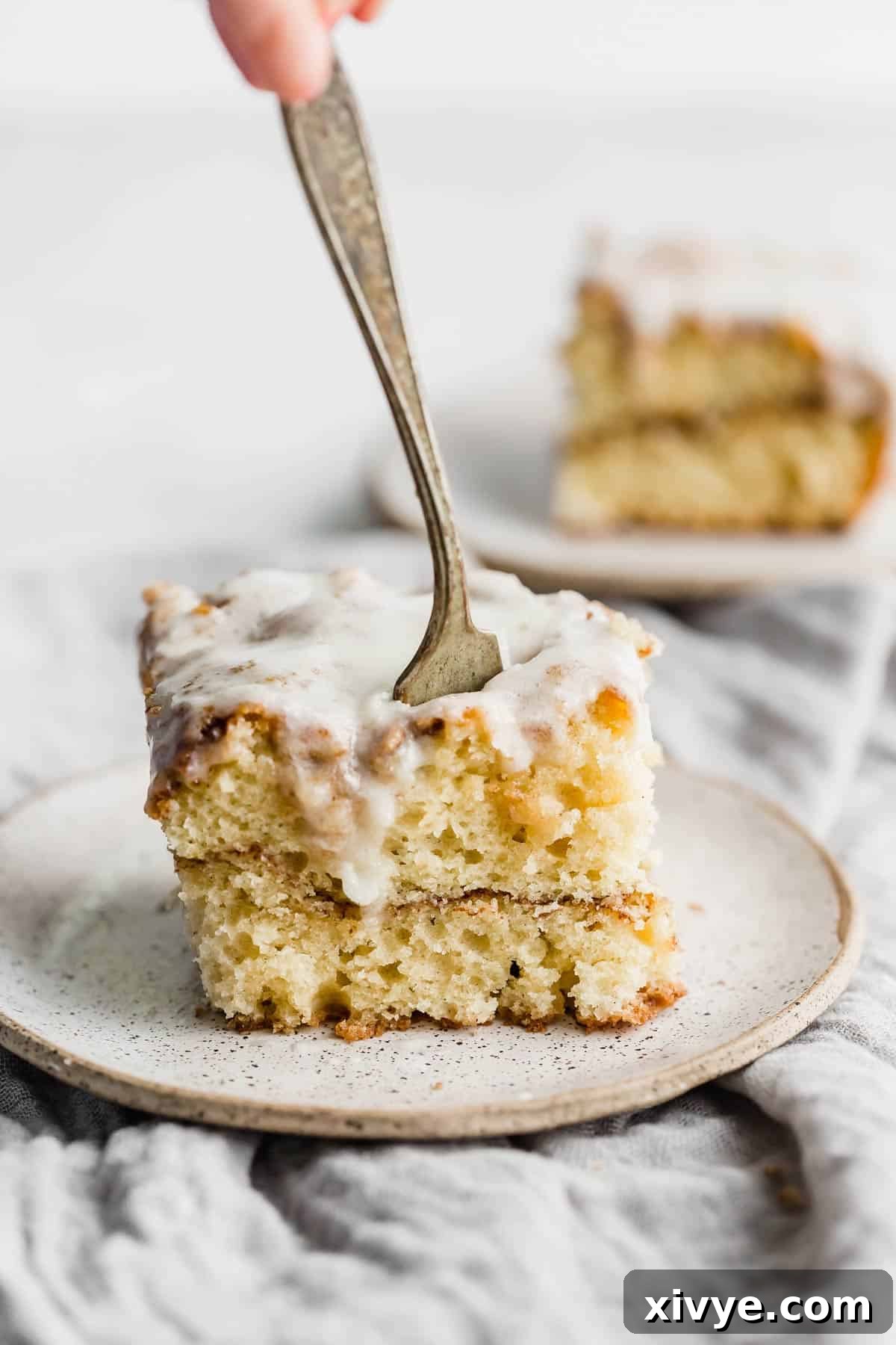 A fork cutting into an easy cinnamon roll cake slice on a white plate, highlighting its soft texture.