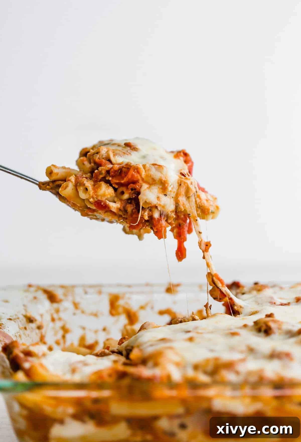 Comforting Sausage Ziti Bake 11 A serving spoon scooping up cheesy baked ziti from a casserole dish, showcasing layers of pasta, sausage sauce, and melted cheese.