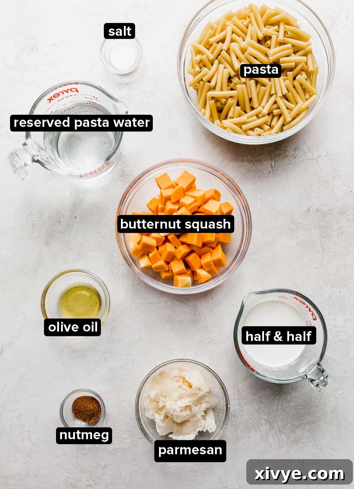 All the fresh ingredients for roasted butternut squash pasta laid out on a clean white surface, showcasing butternut squash, half & half, nutmeg, parmesan, and pasta.