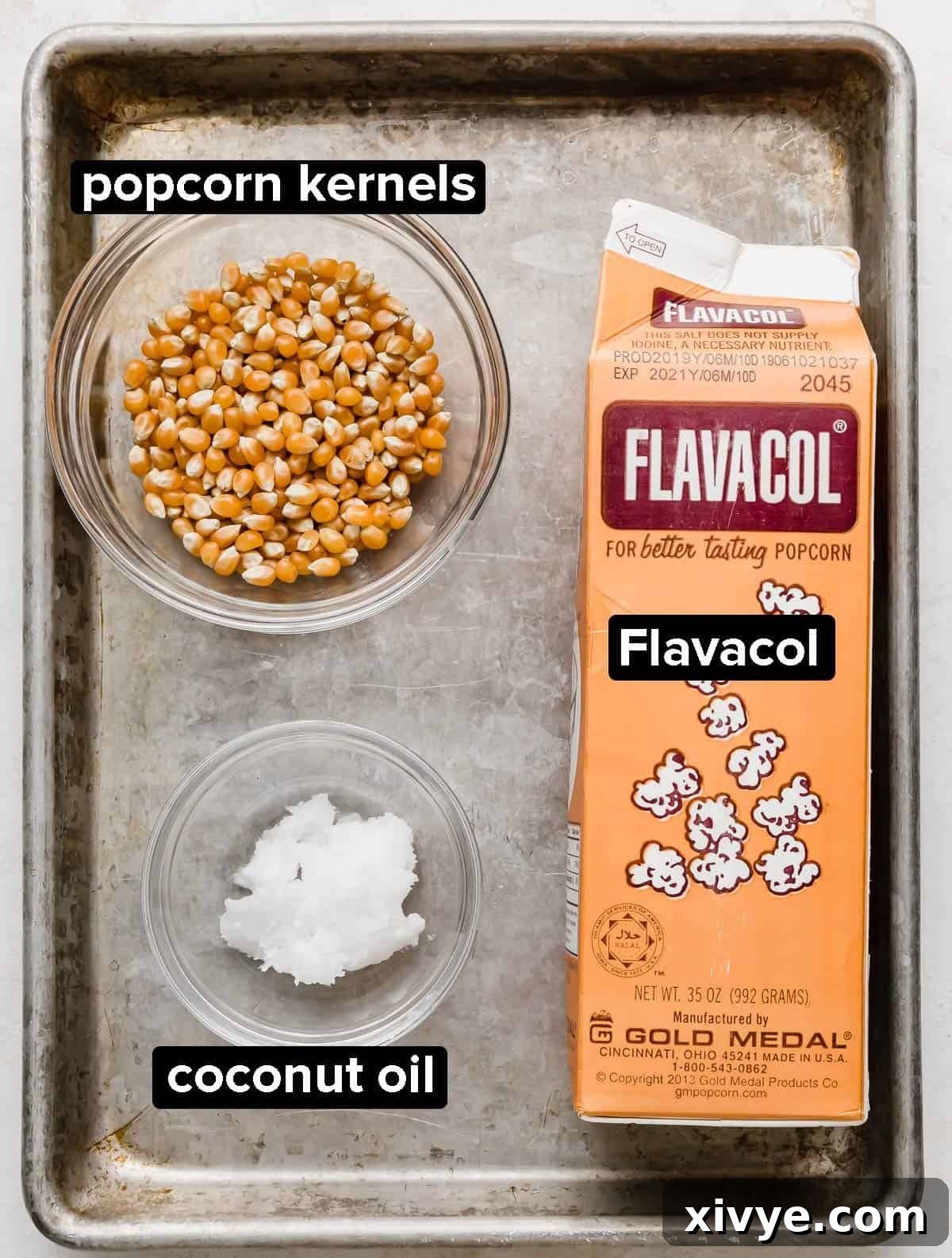 Close-up of three essential ingredients for cinema popcorn: unpopped popcorn kernels, a jar of coconut oil, and a container of Flavacol seasoning, all resting on a gray baking sheet.