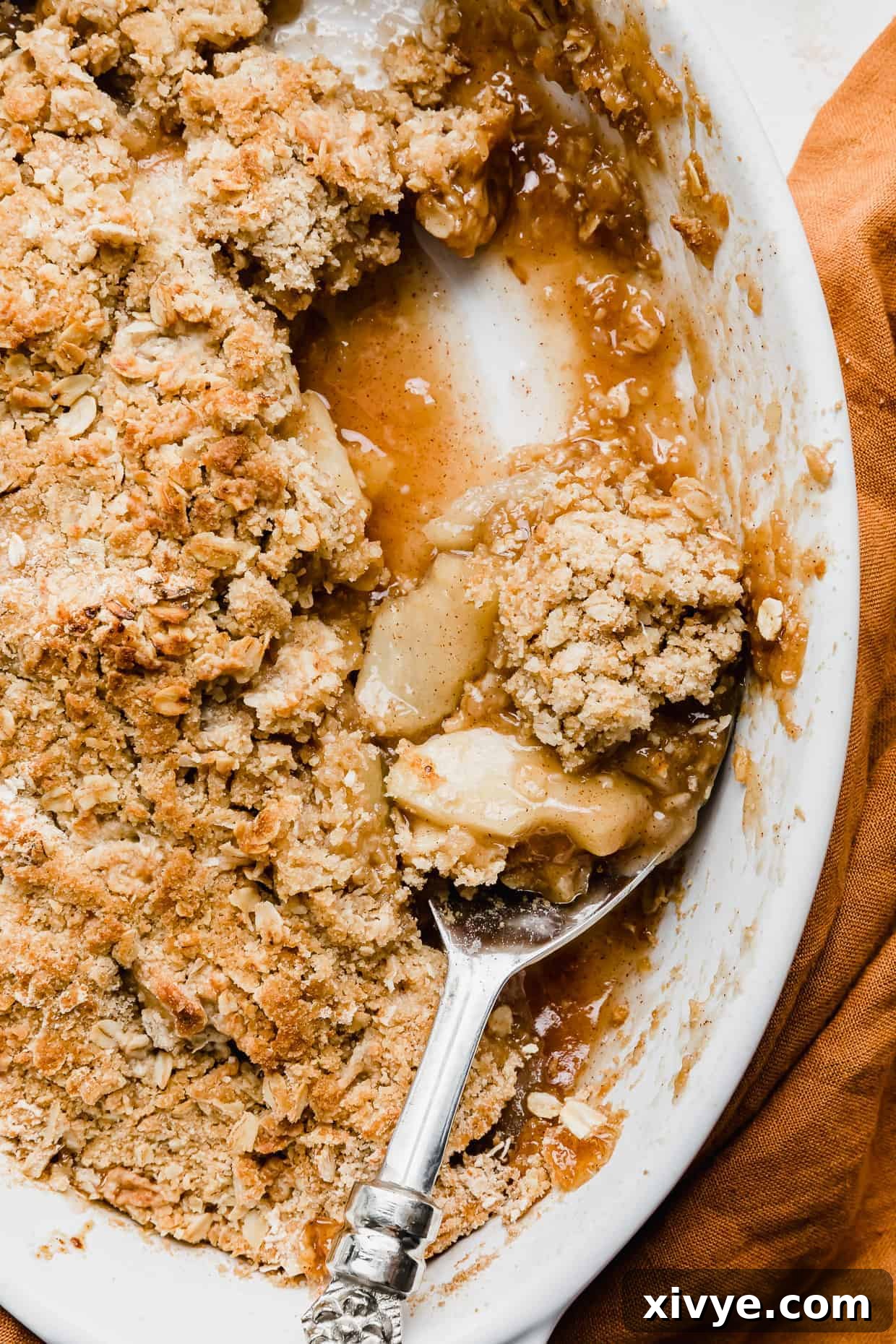 A serving spoon scooping out homemade apple crisp from a white baking dish, showing the buttery oat crumble and soft apple filling.