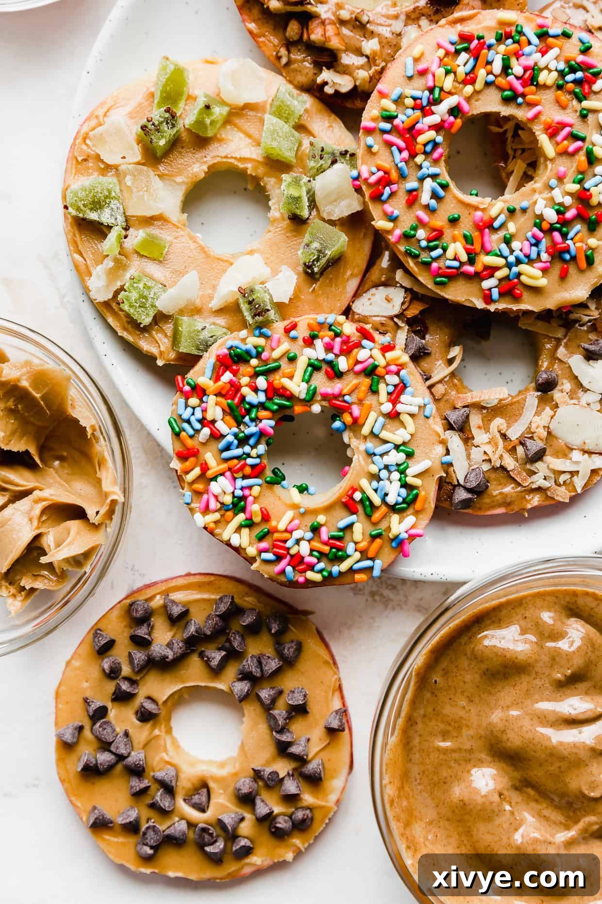 Colorful slices of apples, meticulously cut into round "donuts" and generously topped with creamy peanut butter, vibrant sprinkles, and mini chocolate chips.