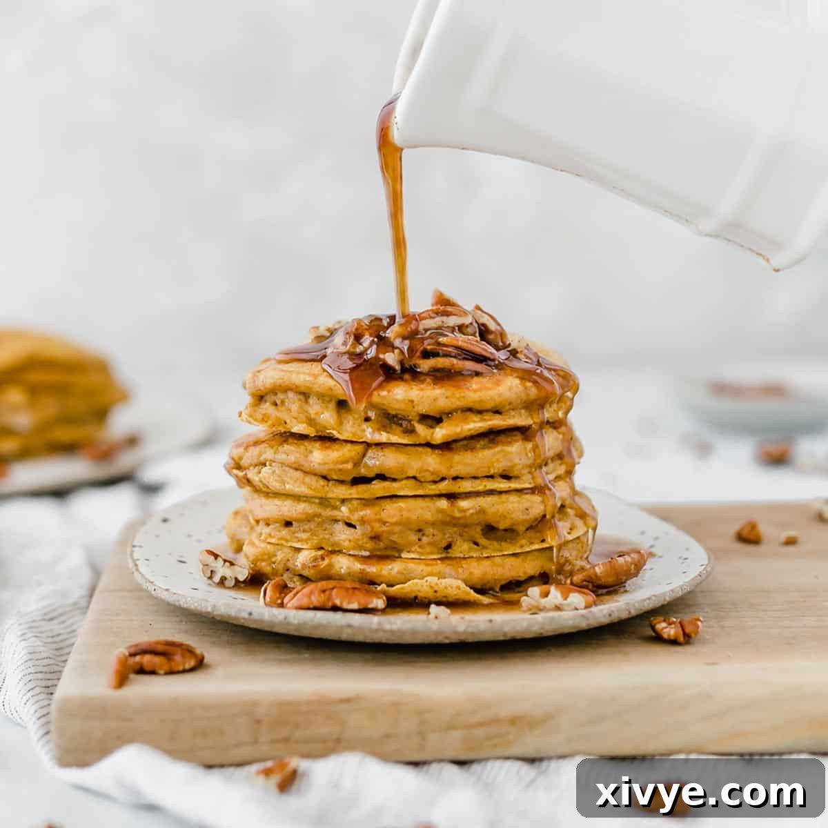 A stack of fluffy pumpkin pancakes topped with pecans, with luscious syrup being poured overtop, promising a delightful breakfast.