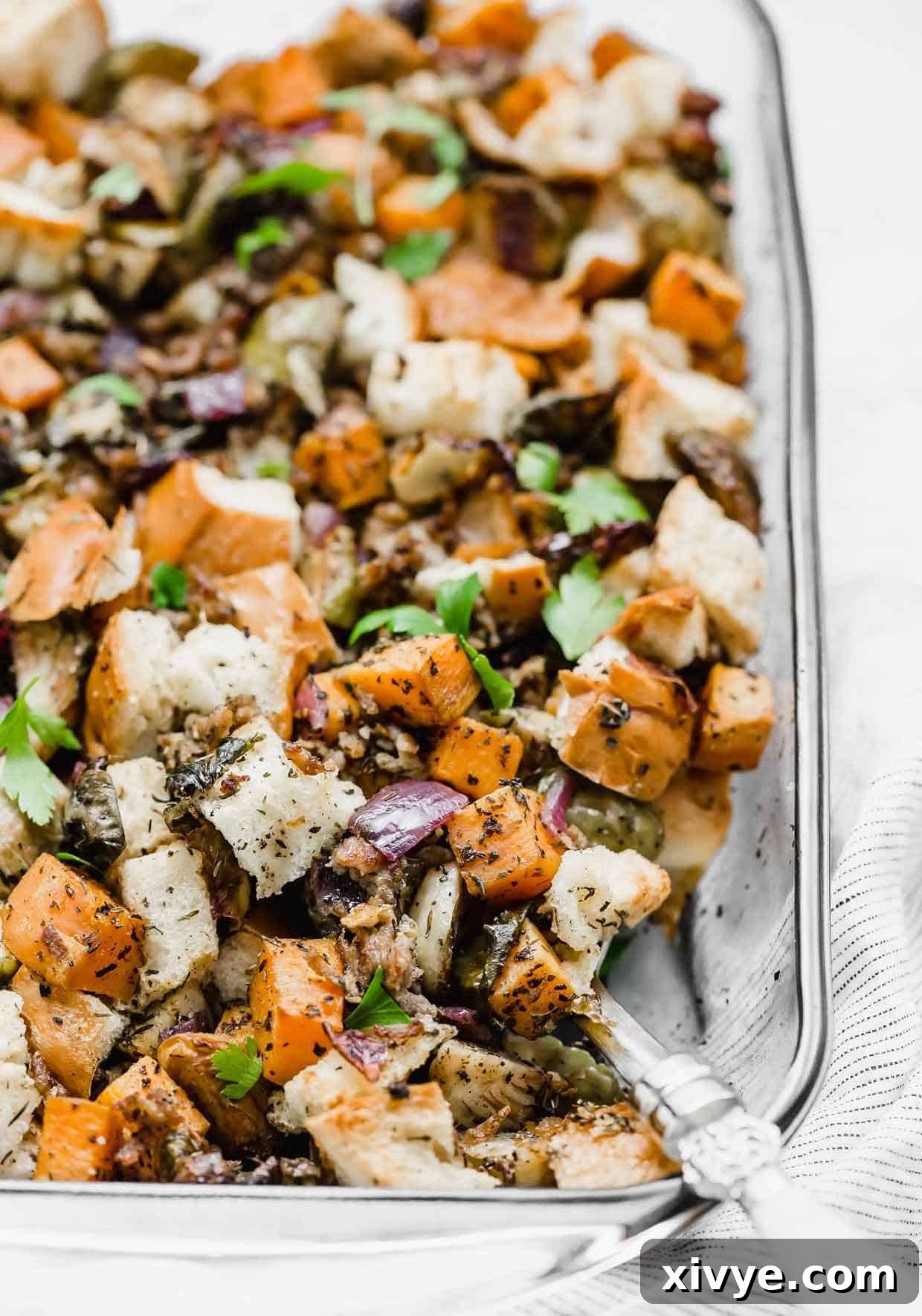 A large casserole dish filled with golden-brown Italian Sausage Stuffing, featuring visible chunks of sweet potato, Brussels sprouts, and French bread, with a serving spoon resting inside.