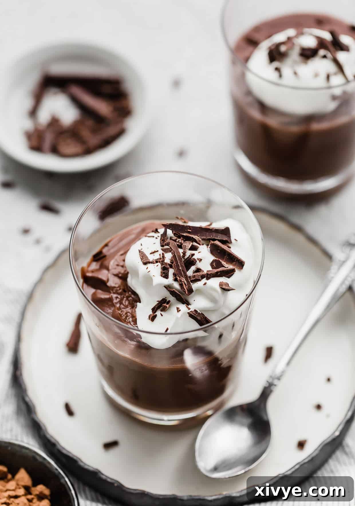 A serving of Homemade Chocolate Pudding in a glass cup, placed on a white plate with a spoon, highlighting its creamy texture.