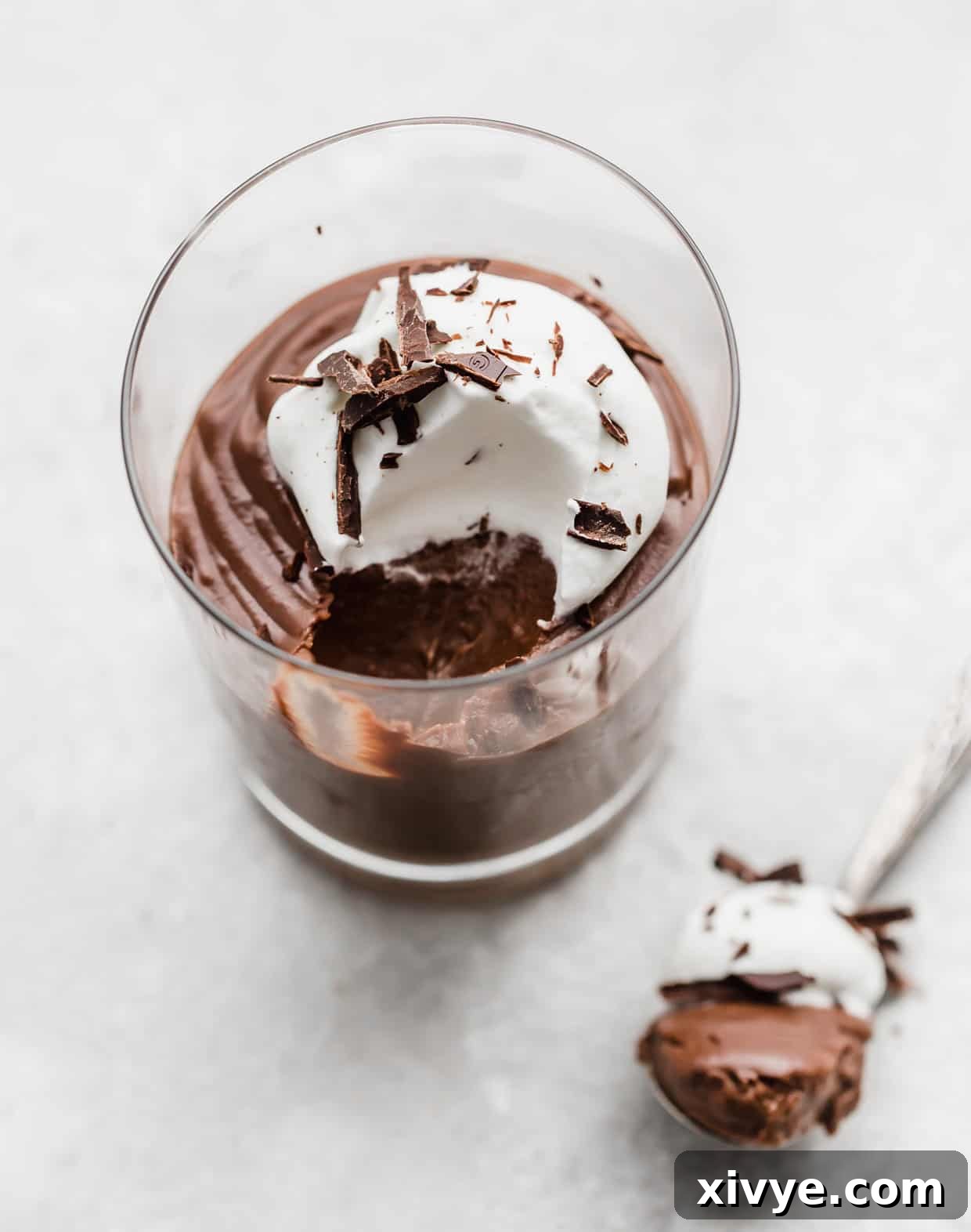 A close-up of dark chocolate pudding in a glass cup, adorned with whipped cream and chocolate shavings, set against a pristine white background.