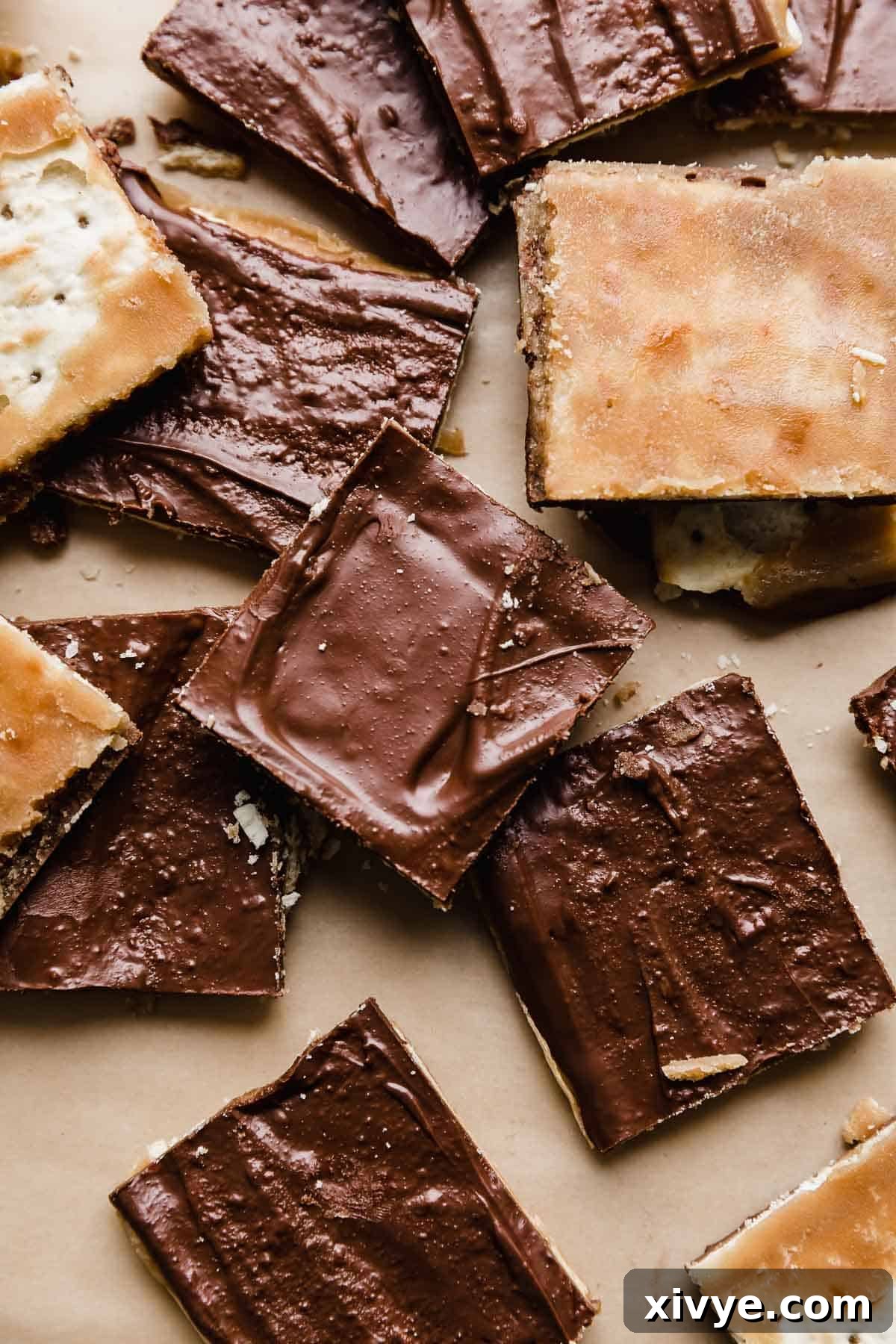 A large sheet of freshly made Christmas Crack, featuring a base of golden-brown saltine crackers, a rich, dark toffee layer, and a top layer of smooth, melted chocolate, set on tan parchment paper. Some pieces are already broken, revealing the crisp layers and tempting texture.