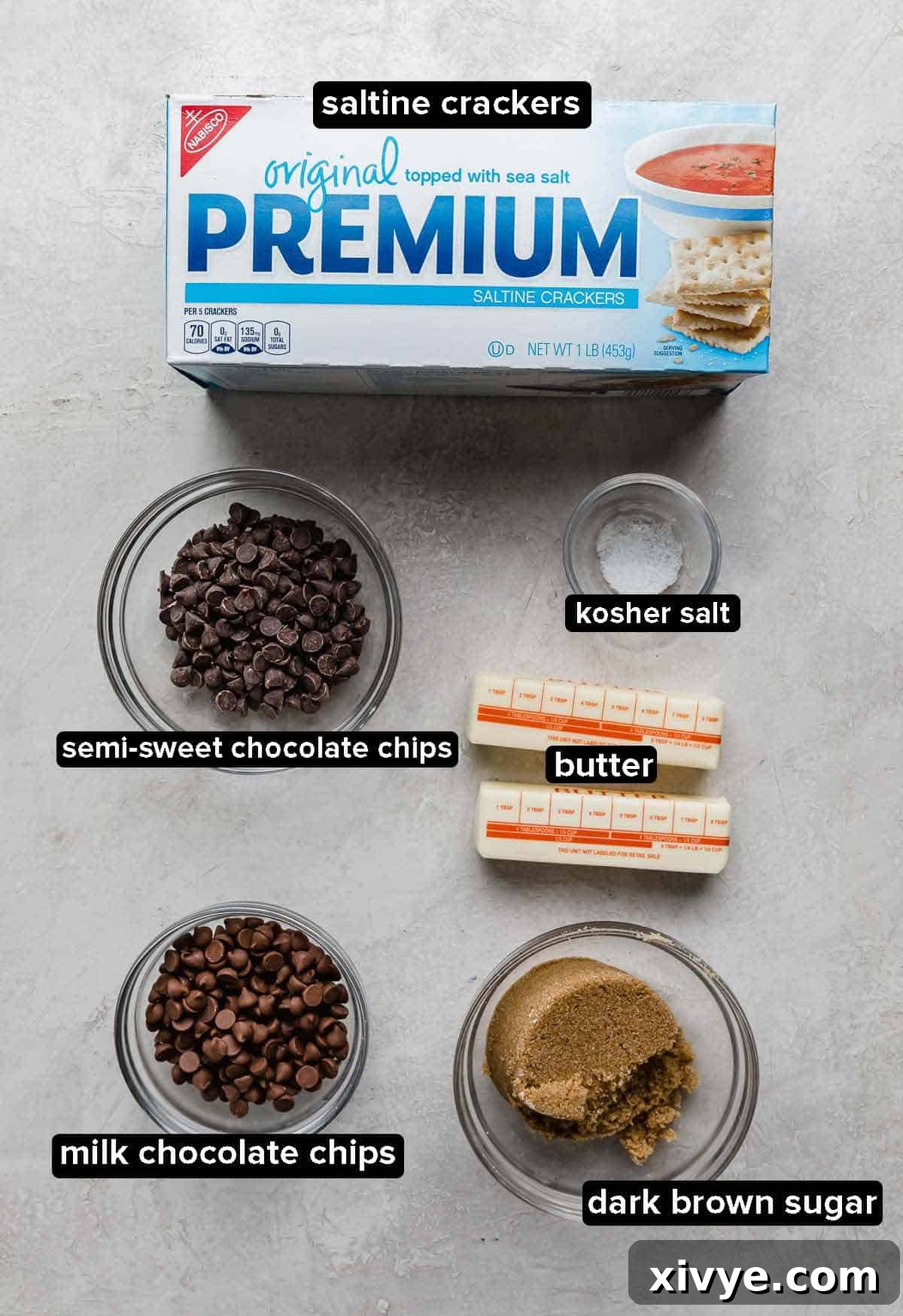 A curated display of Christmas crack ingredients on an elegant gray background, including a box of saltine crackers, a bowl of assorted chocolate chips, a stick of butter, a small dish of kosher salt, and a mound of dark brown sugar, all perfectly arranged and ready for baking.