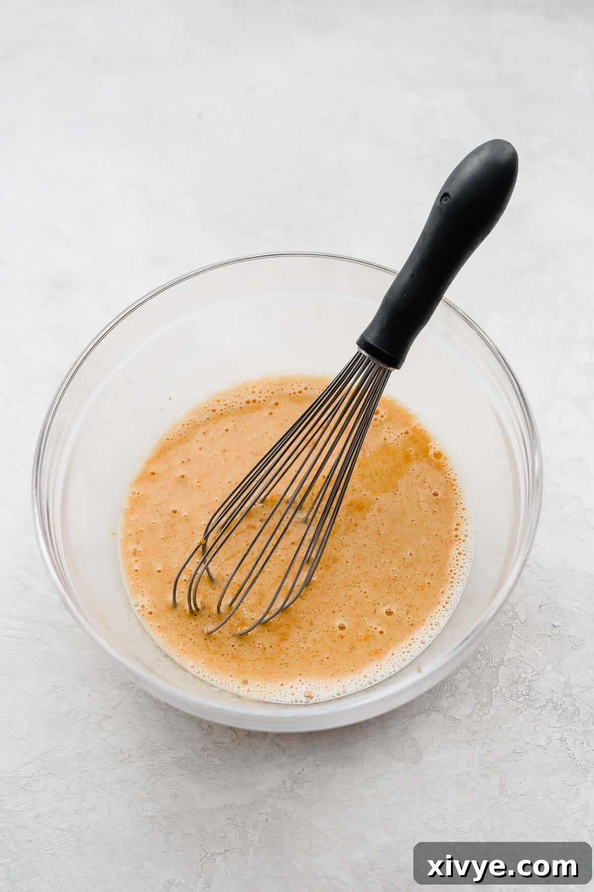 Spiced Pumpkin Oat Muffins 4 A whisk stirring a light orange liquid mixture in a glass bowl, indicating the wet ingredients are being combined for the pumpkin oatmeal muffins.