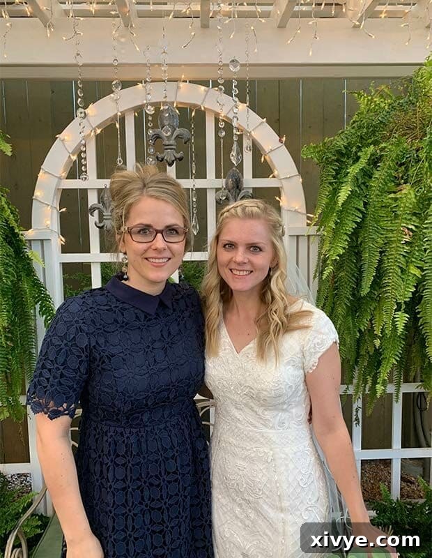 A beautiful wedding photo featuring a bride in a white gown embracing a woman in a navy dress with glasses. They are standing outdoors with a soft focus background, capturing a moment of warmth and celebration.
