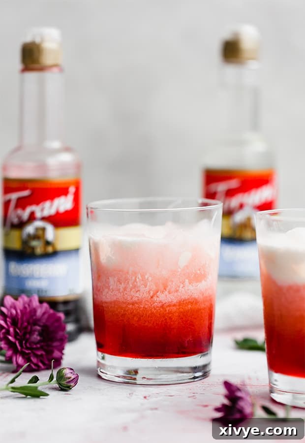 A glass of homemade Italian Cream Soda with raspberry syrup and cream, garnished with a maraschino cherry. The drink shows beautiful layers.