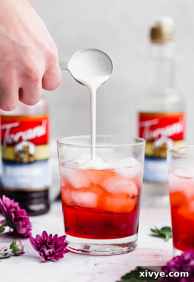 Heavy cream being slowly poured into a glass cup containing club soda and syrup, creating beautiful swirls as it mixes into the Italian cream soda.