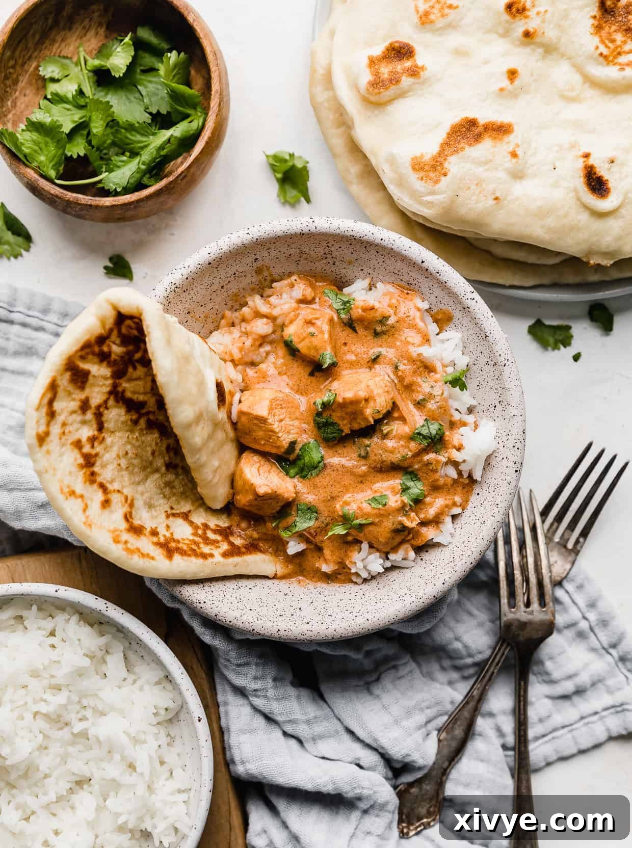 A bowl of Indian Butter Chicken Recipe surrounded by naan bread and cilantro.