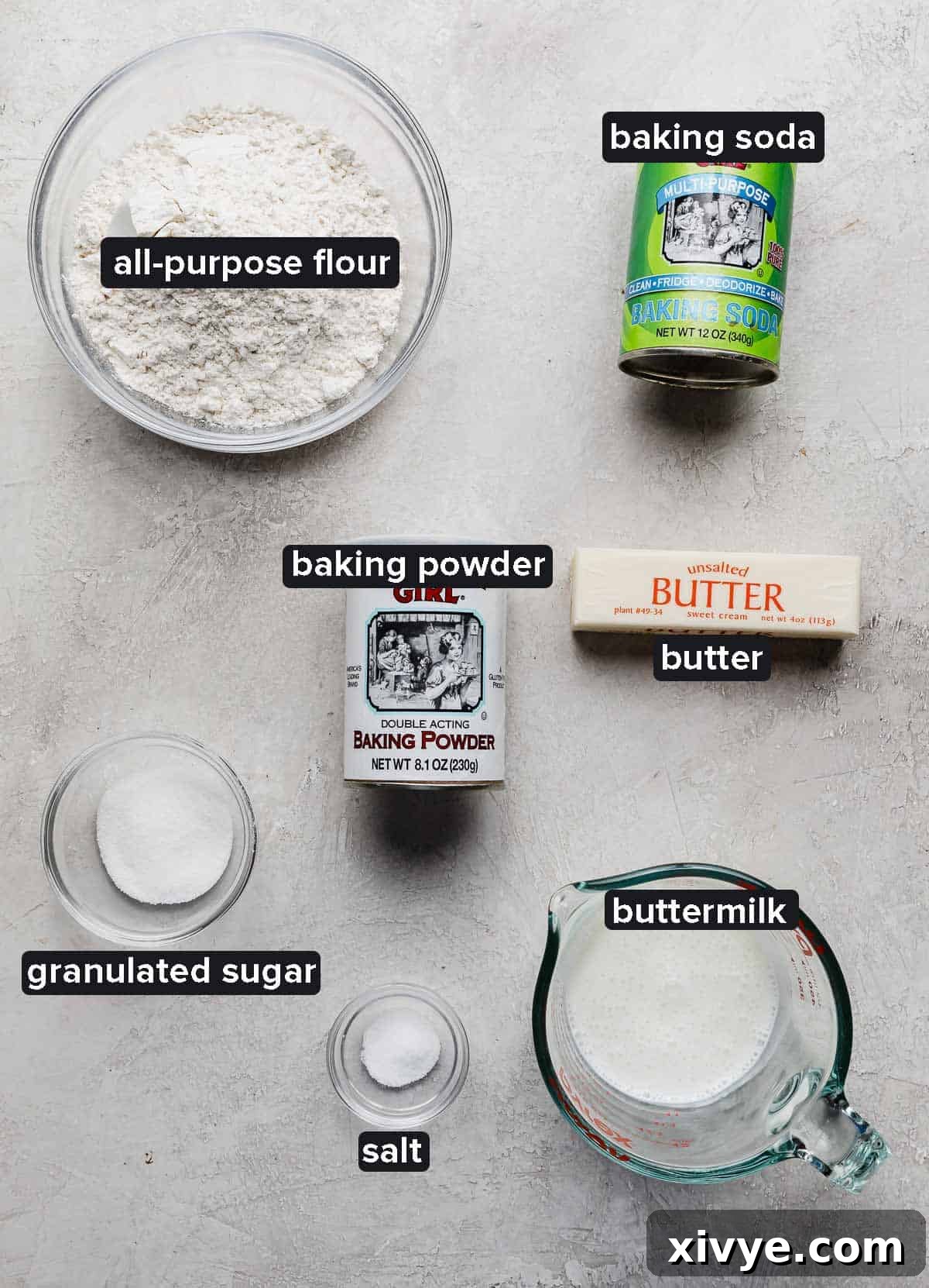 Buttermilk biscuit ingredients portioned into glass bowls on a light gray textured background: flour, buttermilk, baking powder and soda, salt, sugar, and butter.