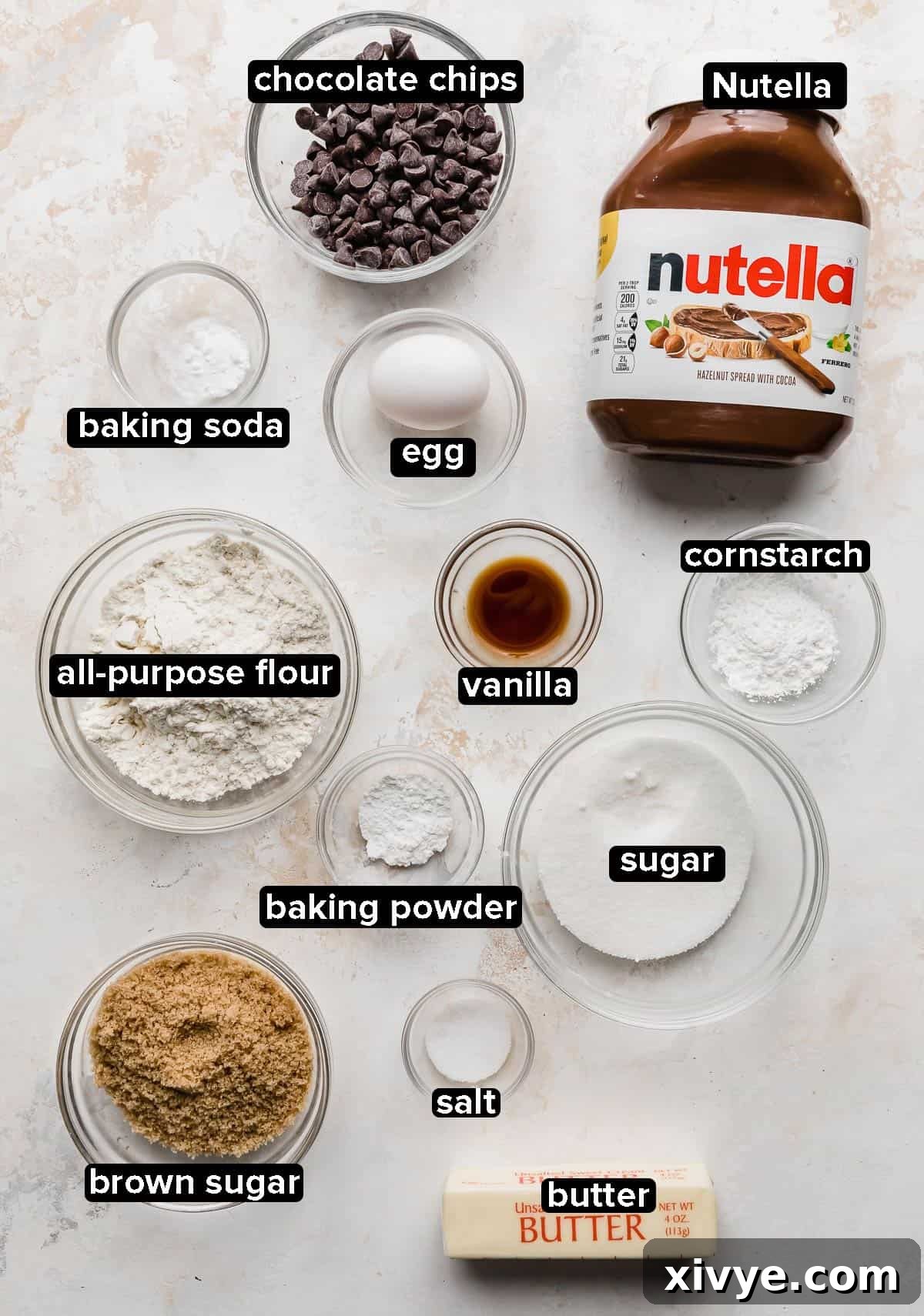 Ingredients used to make Nutella Stuffed Nutella Cookies portioned into glass bowls on a white background, including butter, sugars, Nutella, egg, vanilla, cornstarch, and chocolate chips.