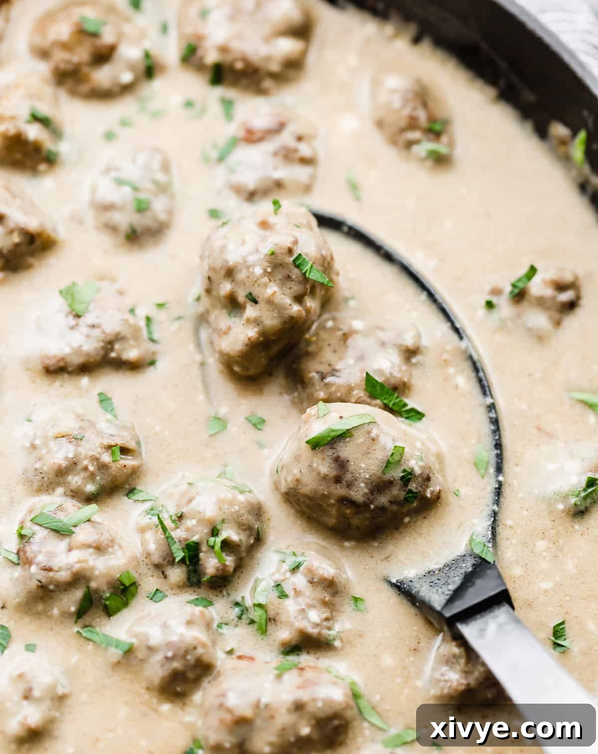 A large spoon scooping into a skillet full of Swedish Meatballs and tan colored sauce.