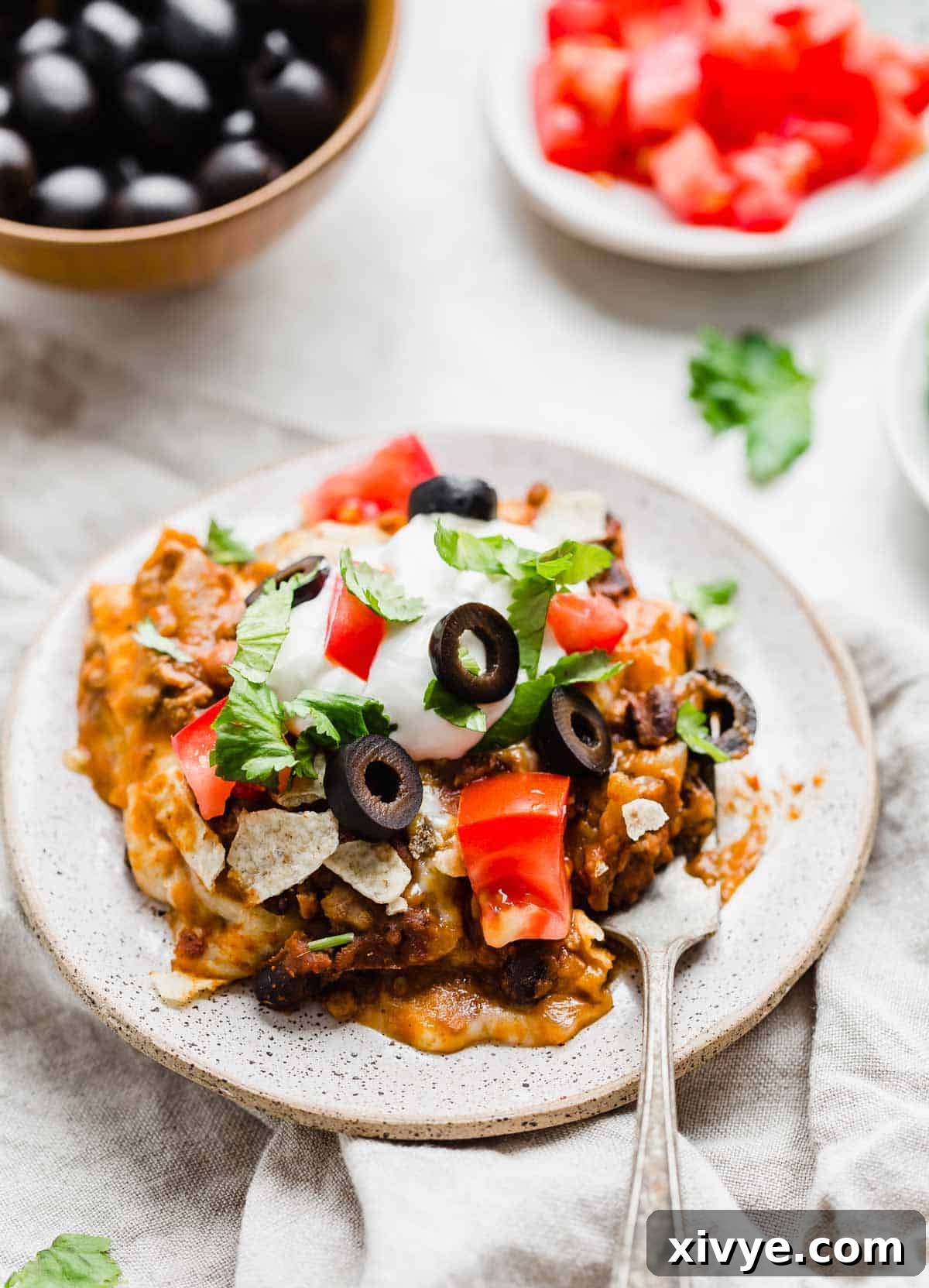 A slice of Mexican Lasagna on a plate topped with fresh tomatoes, olive, lettuce, and sour cream.