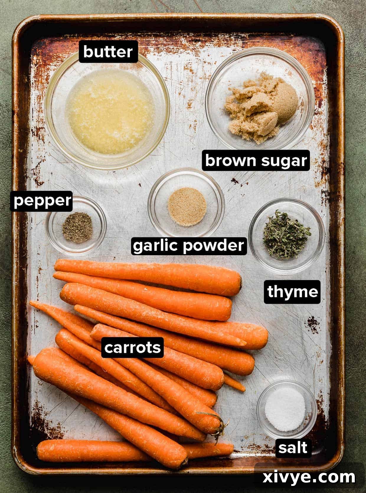 All ingredients for brown sugar roasted carrots laid out on a baking sheet: fresh carrots, butter, brown sugar, thyme, garlic powder, salt, and pepper.