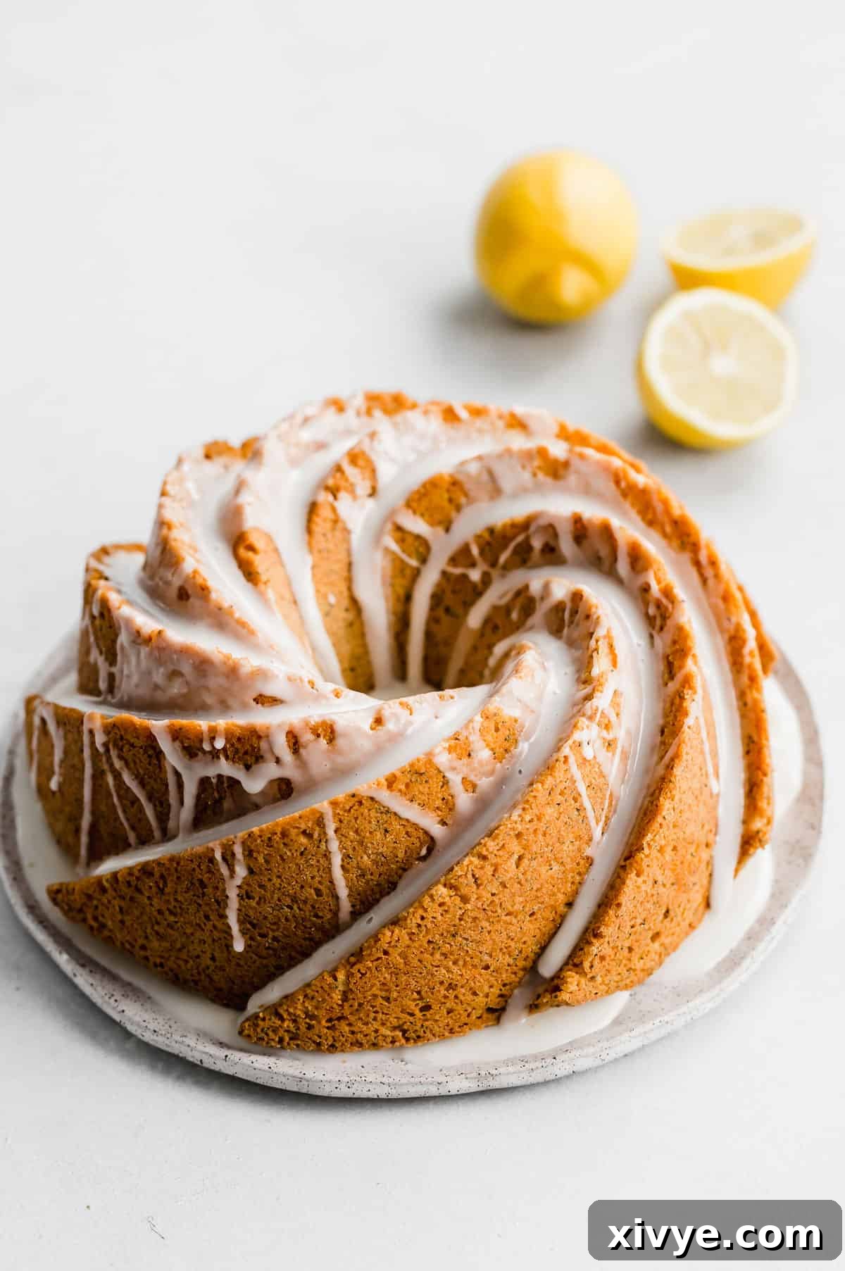 A Lemon Poppy Seed Bundt Cake topped with a white glaze on a white plate on a white background, ready to be served.