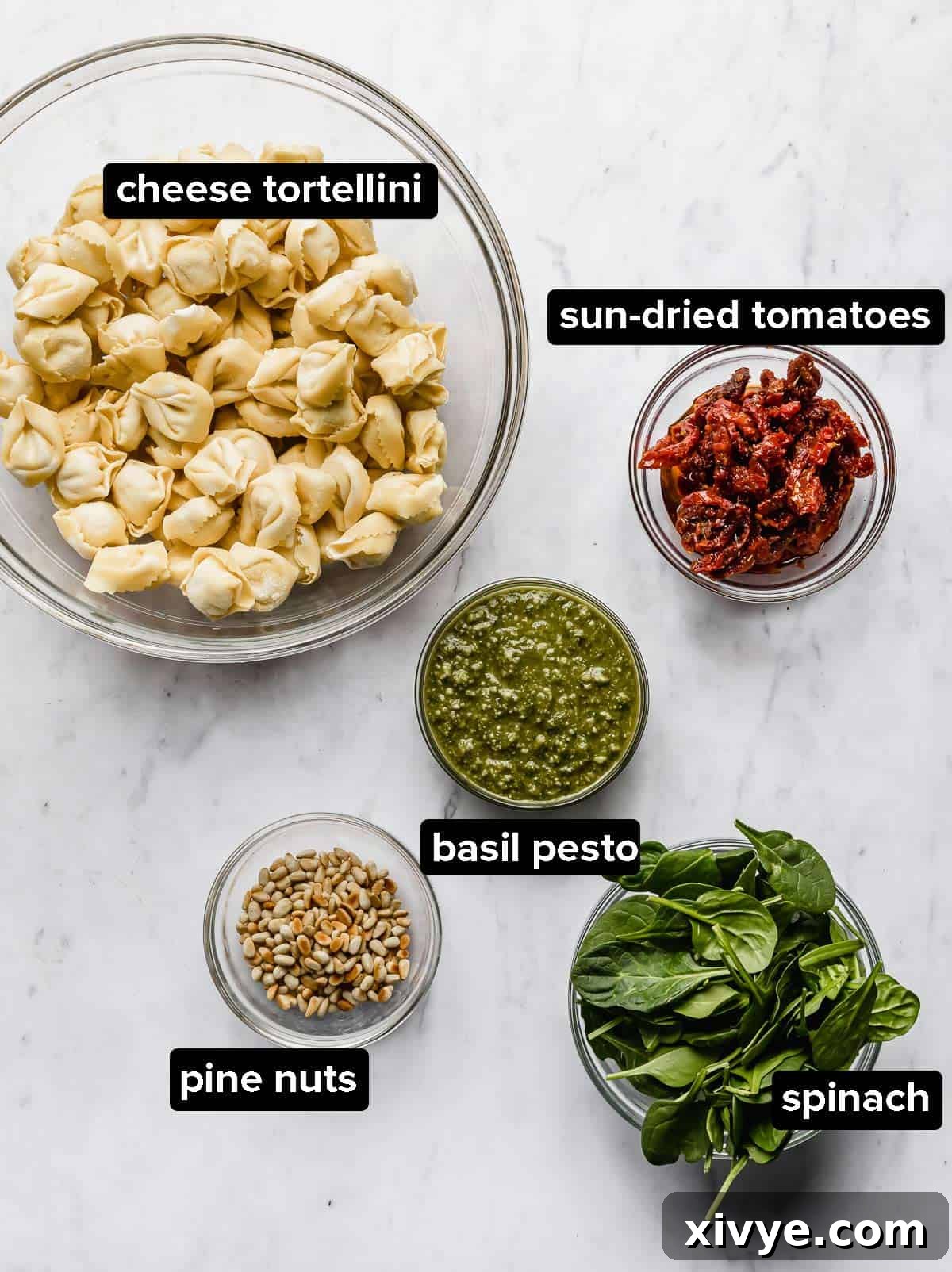 Ingredients used to make Pesto Tortellini Pasta Salad, portioned into glass bowls: tortellini, pesto, sun-dried tomatoes, pine nuts, and optional spinach.