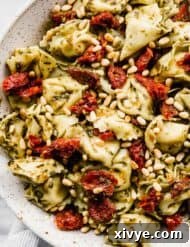 A close up photo of Pesto Tortellini Pasta Salad tossed with sun-dried tomatoes and pine nuts in a white bowl.