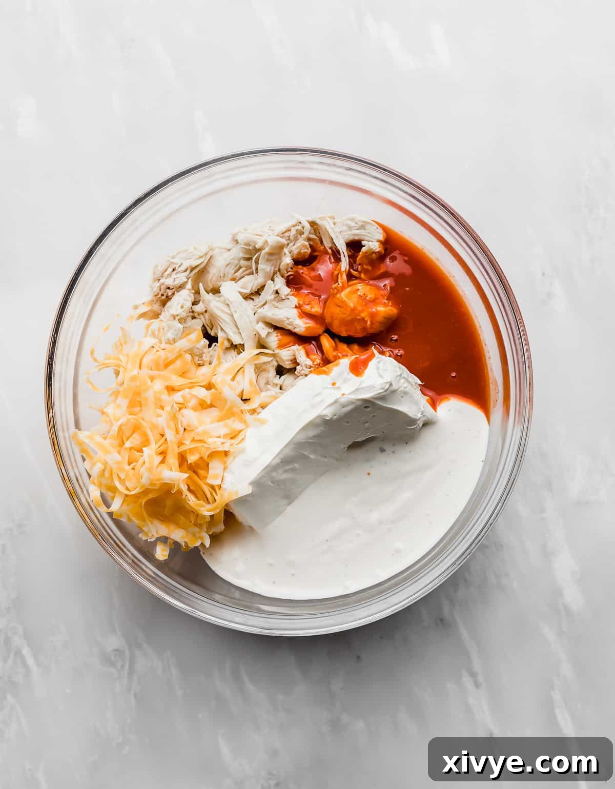 The Ultimate Buffalo Chicken Dip 4 A glass bowl filled with shredded chicken, softened cream cheese, vibrant buffalo sauce, creamy ranch dressing, and shredded cheese, ready to be mixed for the perfect dip.