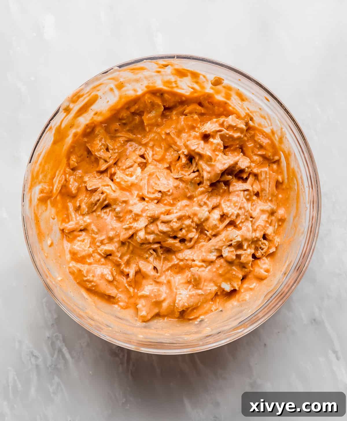 The Ultimate Buffalo Chicken Dip 5 A vibrant orange Buffalo Chicken Dip mixture, perfectly combined and smooth, resting in a glass bowl on a pristine white marble background.