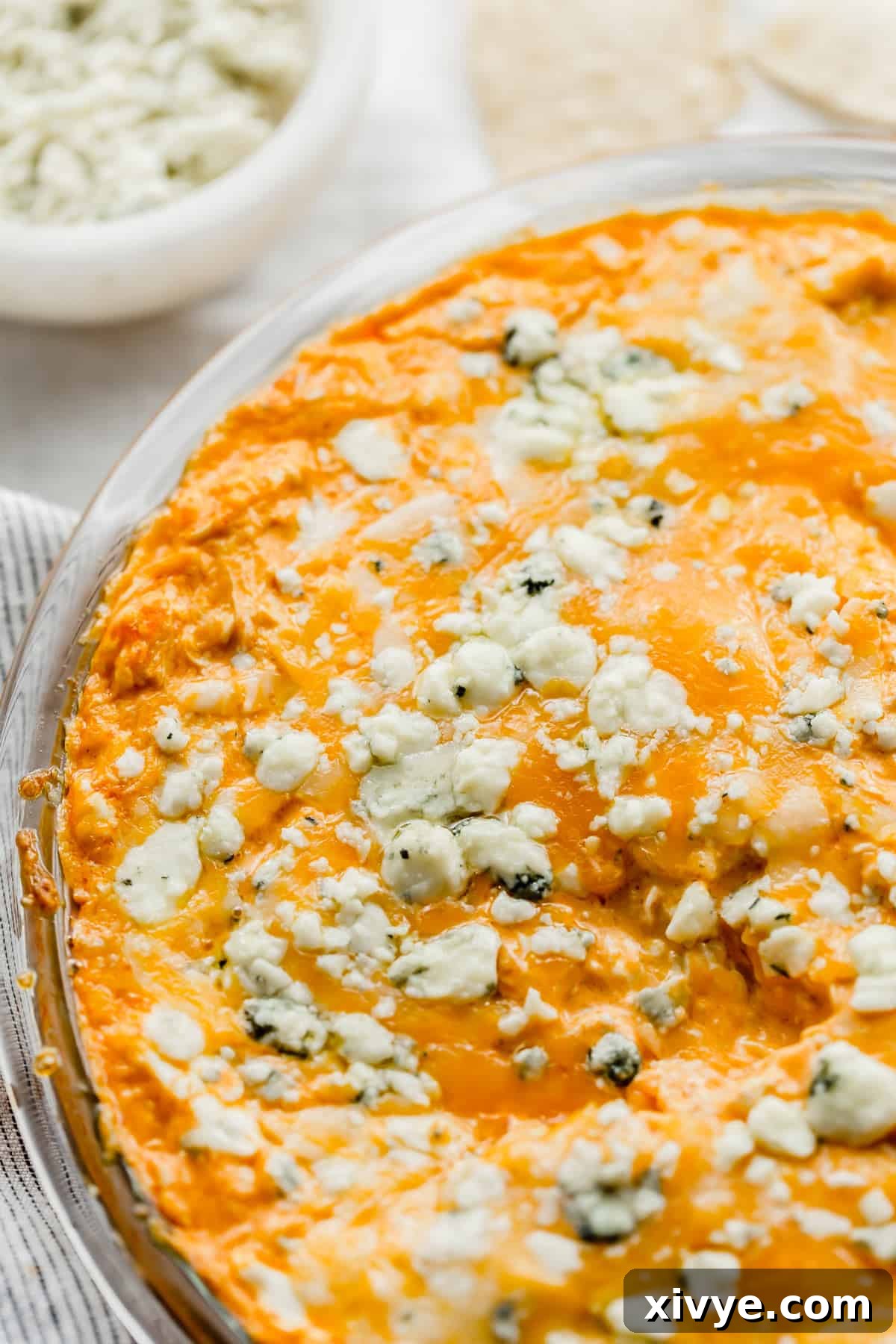 The Ultimate Buffalo Chicken Dip 9 A top-down view of a perfectly baked Buffalo Chicken Dip, generously topped with blue cheese crumbles, served in a round pie plate, showcasing its golden bubbly edges.