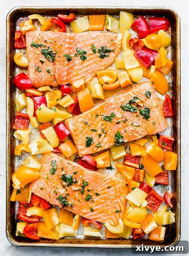 Overhead photo of 3 salmon pieces on a baking sheet surrounded by yellow, orange, and red bell pepper pieces, cooked.
