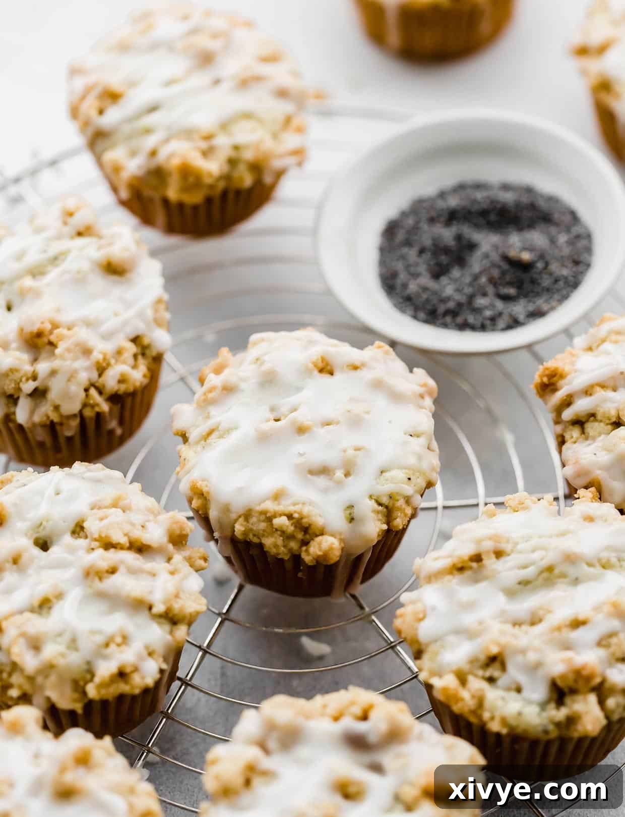 Zesty and Tender Lemon Poppy Seed Muffins 9 Glazed and crumb topped Lemon Poppy Seed Muffins on a wire rack with a bowl of poppy seeds in the background.