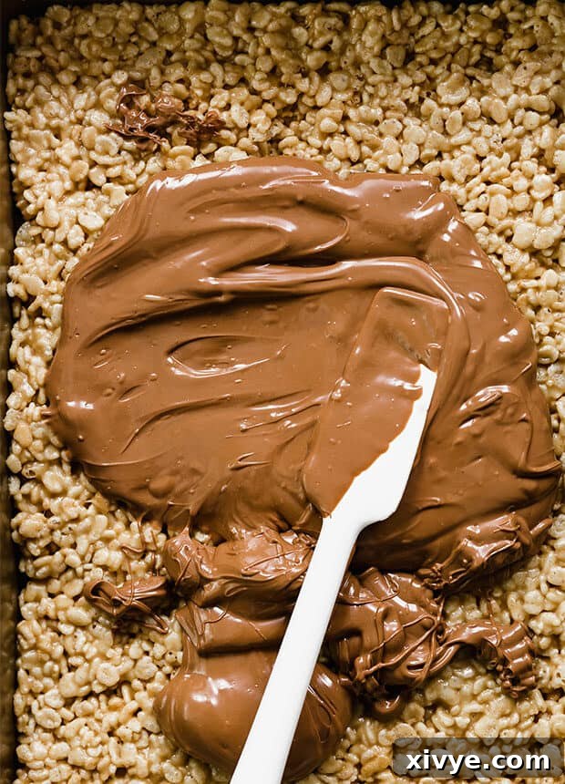 A spatula spreading melted chocolate and butterscotch topping over the Scotcharoo base in a casserole dish.