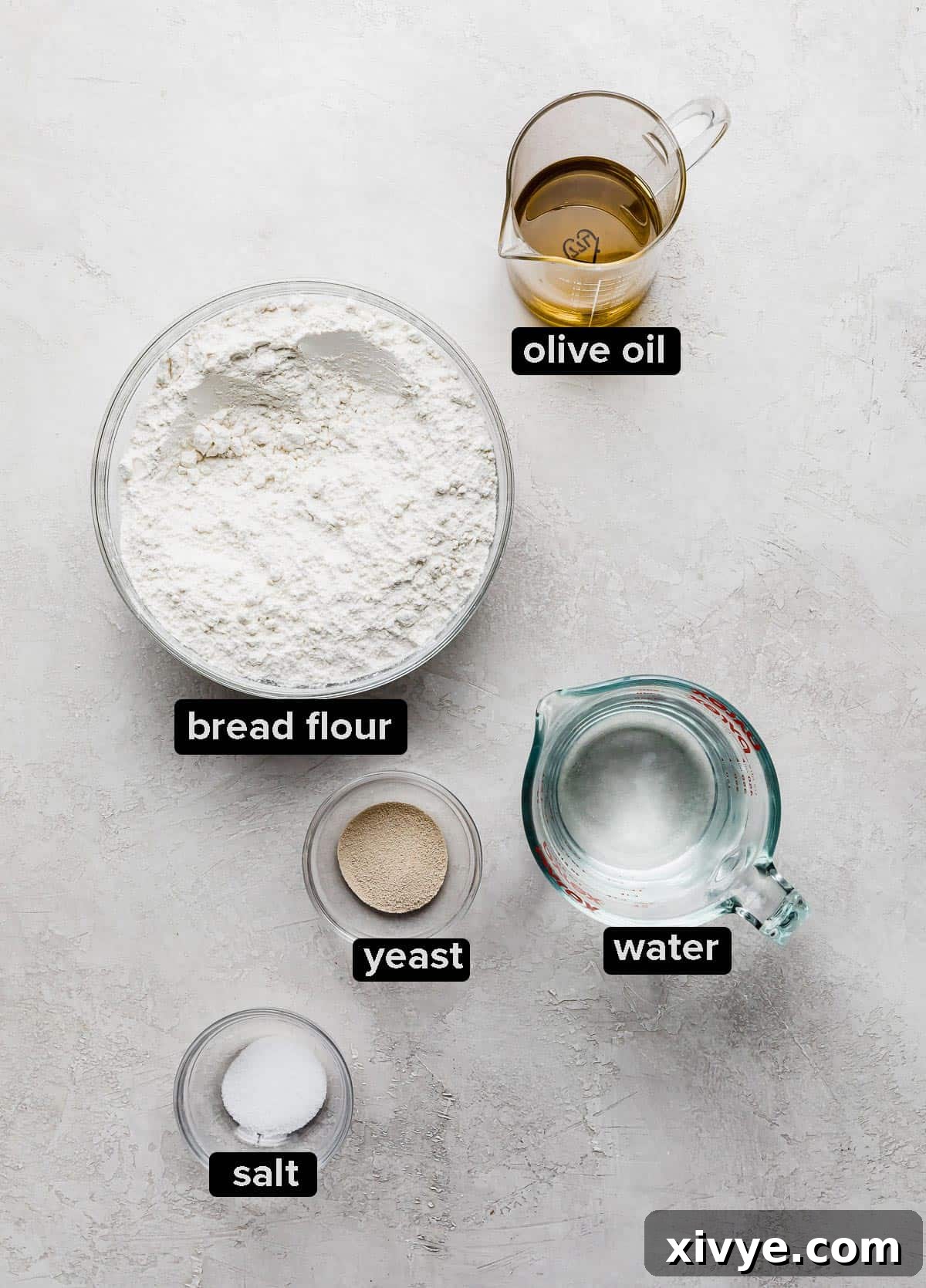 All the essential Focaccia Bread dough ingredients, including bread flour, salt, instant yeast, olive oil, and lukewarm water, neatly arranged on a textured white background.