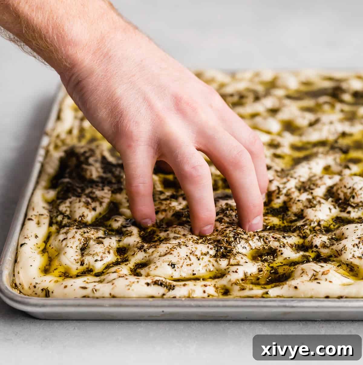 A hand pressing five fingers down into focaccia bread dough topped with herb oil, creating the characteristic dimples before baking.