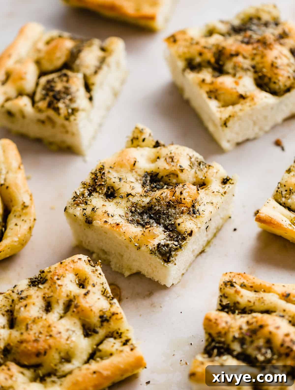 Focaccia Bread squares topped with fresh herbs, elegantly arranged on a light beige background, ready to be served.
