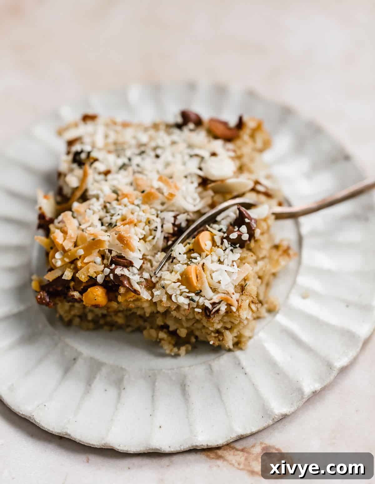 A square slice of Breakfast Baked Oatmeal Cake topped with butterscotch chips, coconut, hemp hearts, and chocolate chips, showcasing its rich texture and diverse toppings.
