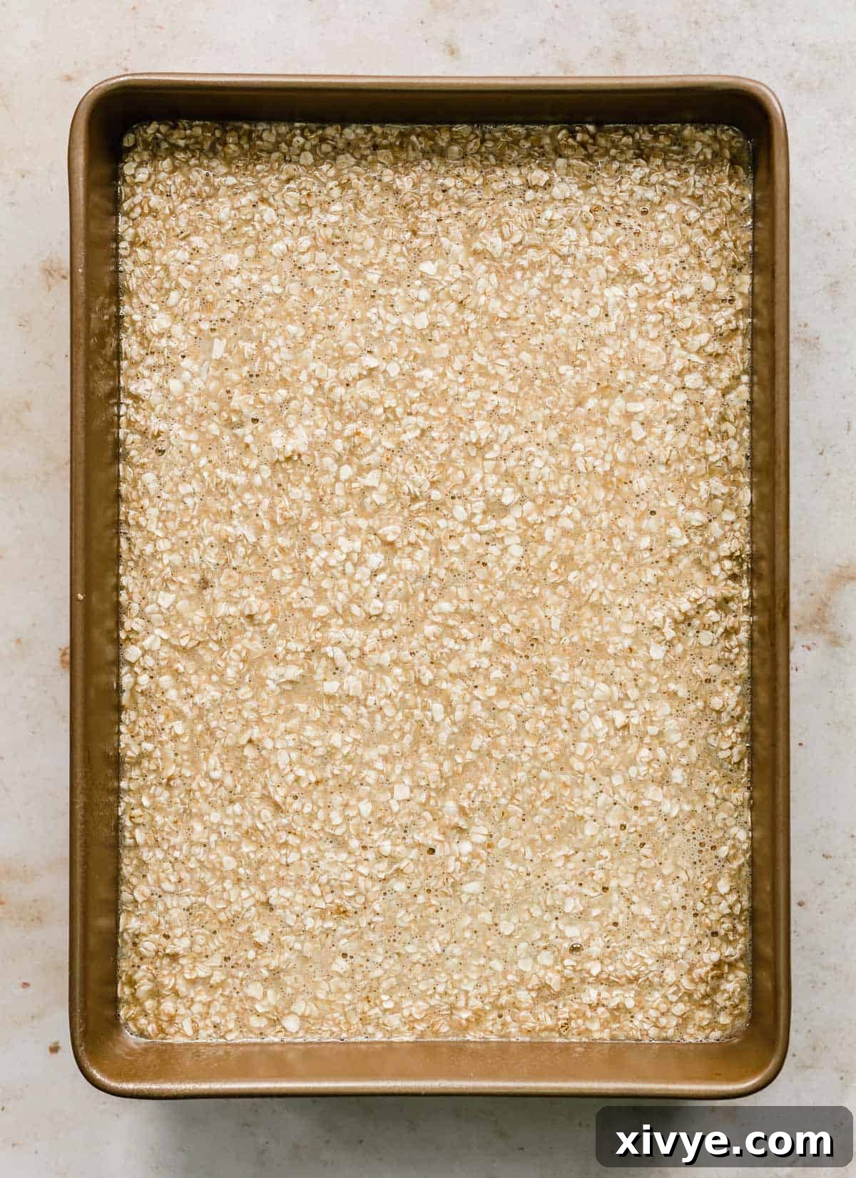 A bronze rectangular baking dish full of oatmeal cake batter, smoothed into an even layer, ready for the toppings and oven.