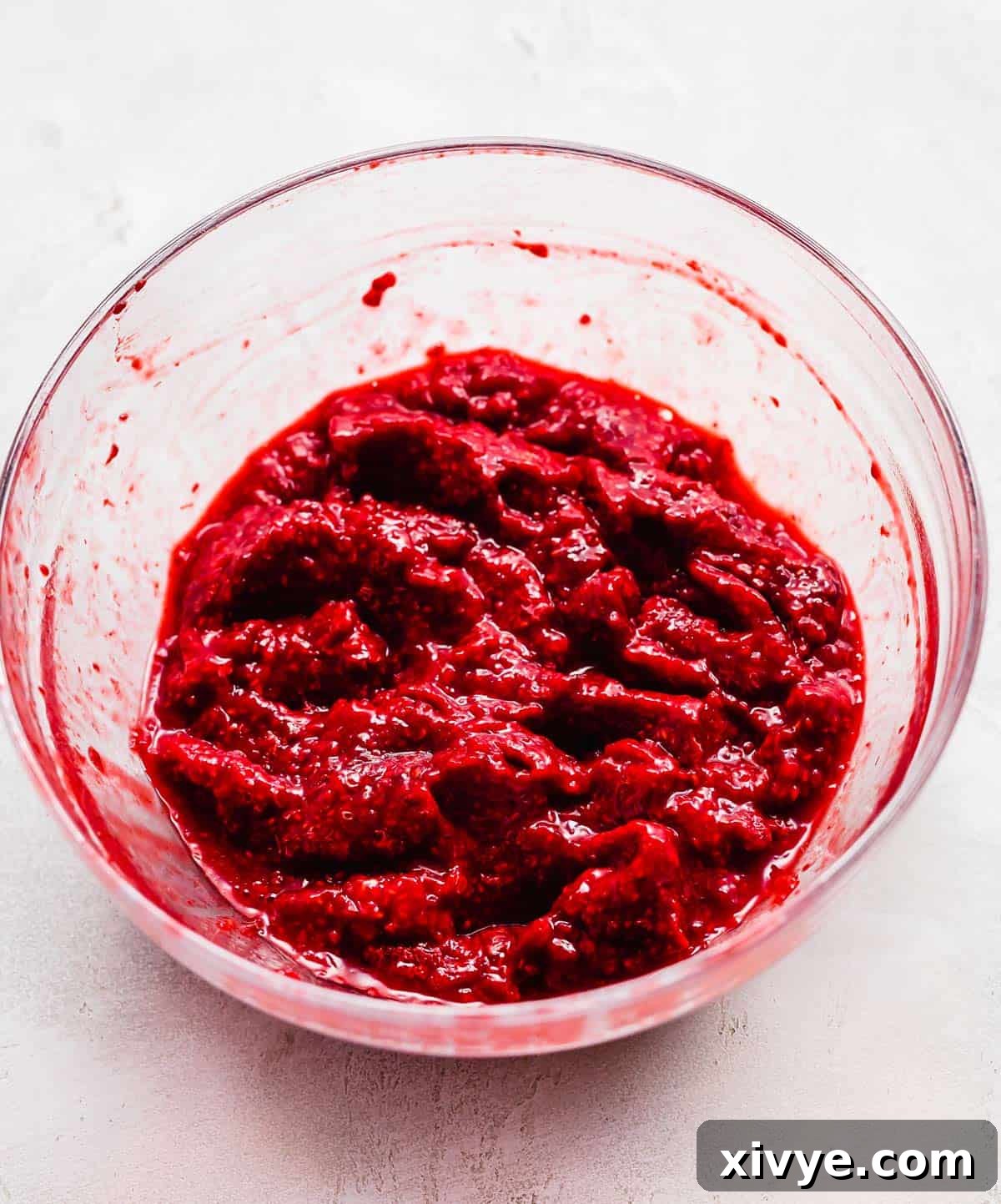 No-Fuss Raspberry Freezer Jam 5 Mashed frozen raspberries to make freezer jam in a glass bowl on a white background.
